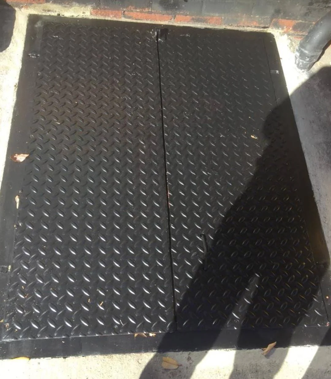 A black diamond-plate metal cover on the ground, divided into two sections, with some leaves and debris around it and a piece of a metal pole visible on the right side.