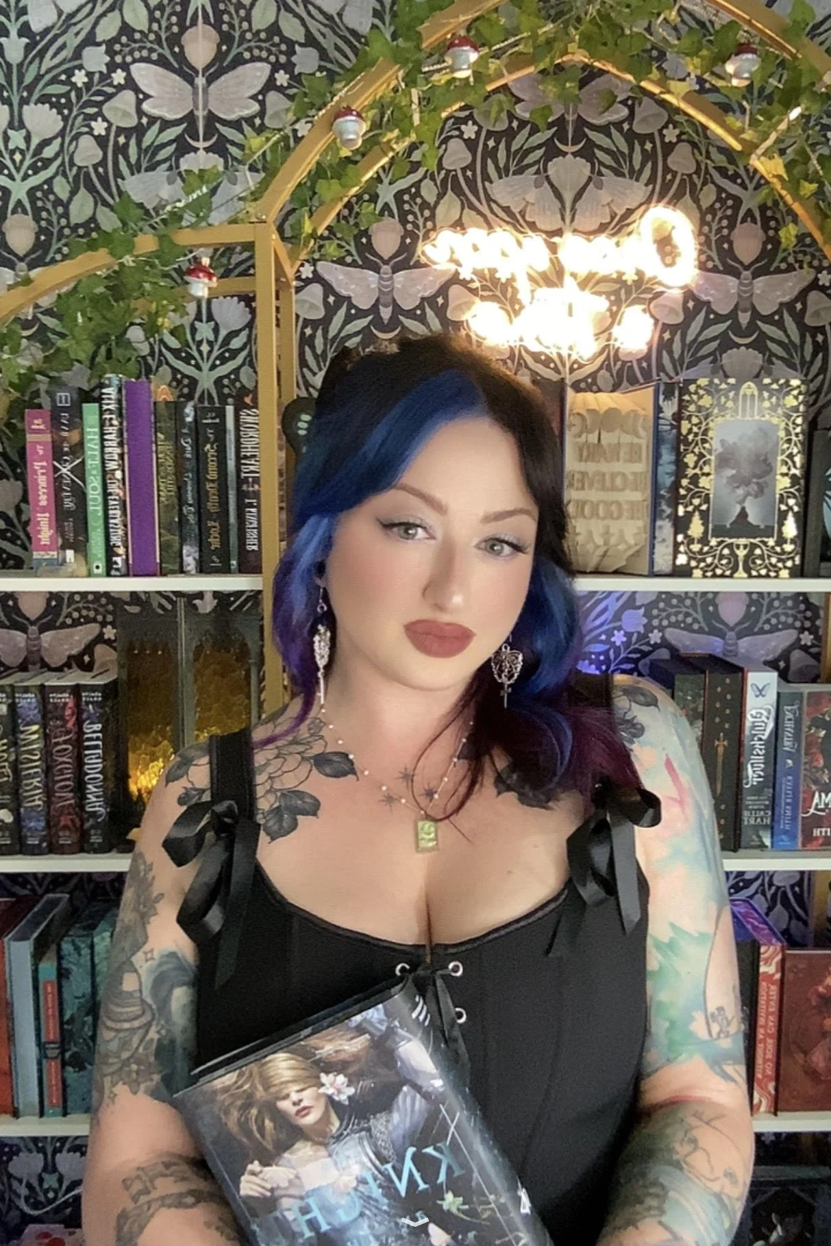 A woman with black and blue hair, tattoos on her arms and shoulders, wearing a black top with ribbons, holding a book with a photo of a woman on the cover, sitting in front of a bookshelf, with a decorative wallpaper background, a chandelier, and greenery.