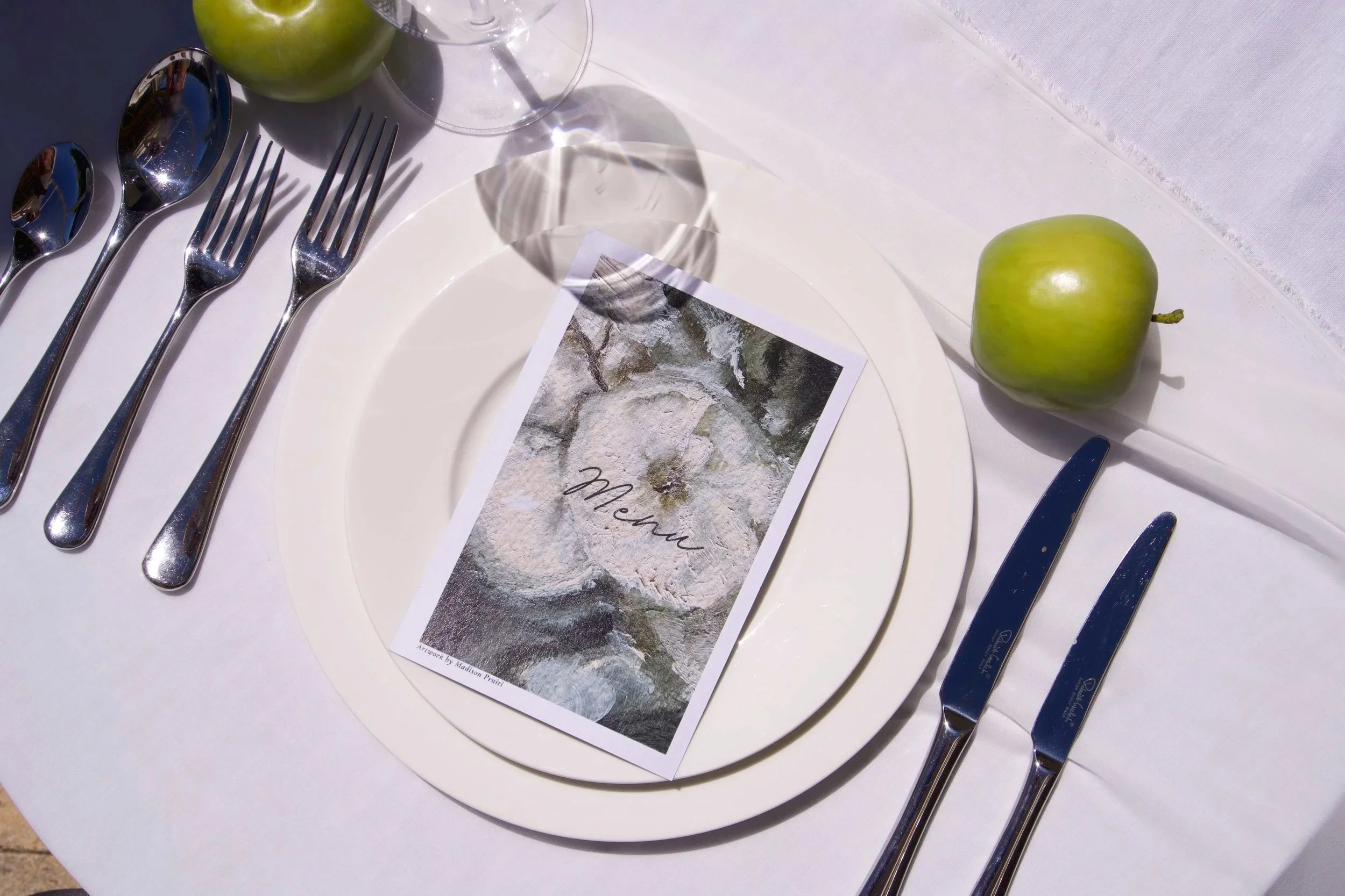 A place setting at a table with white plates, silver cutlery, a green apple, a menu, and wine glasses on a white tablecloth.