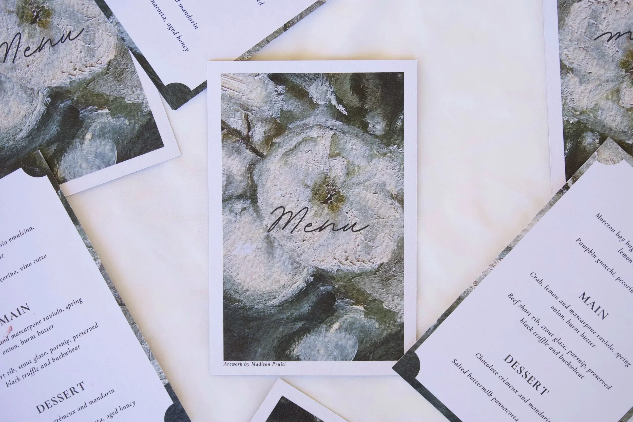 Multiple restaurant menu cards with black and white text, featuring dishes and descriptions, arranged on a white surface. One card has an artistic watercolor-style image of tree rings with the word "Menu" written on it.