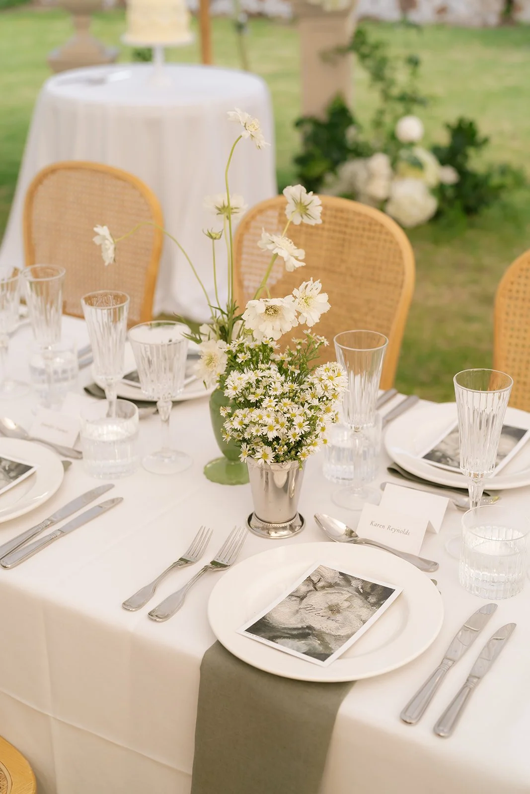 Elegant outdoor table setting with white tablecloth, floral centerpiece with white flowers, wine glasses, water glasses, and silverware, and place cards with handwritten names, on a grassy area.