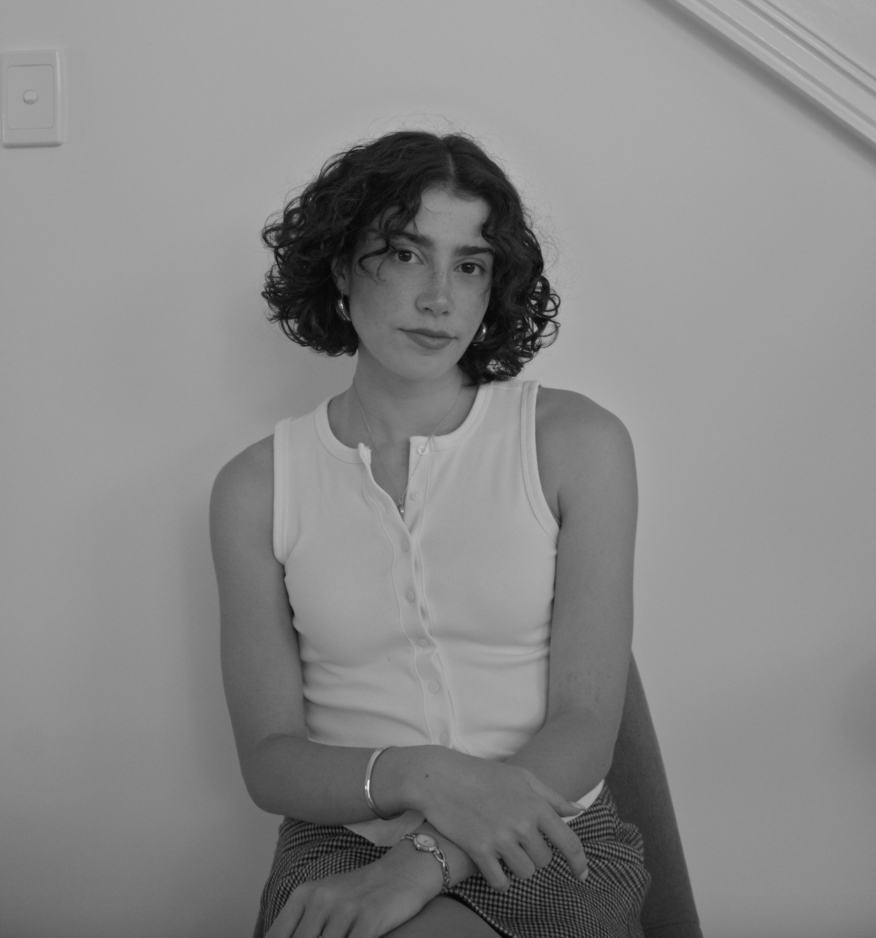 A woman with curly hair wearing a sleeveless white top, sitting on a chair against a plain wall, looking at the camera with a neutral expression in black and white photograph.