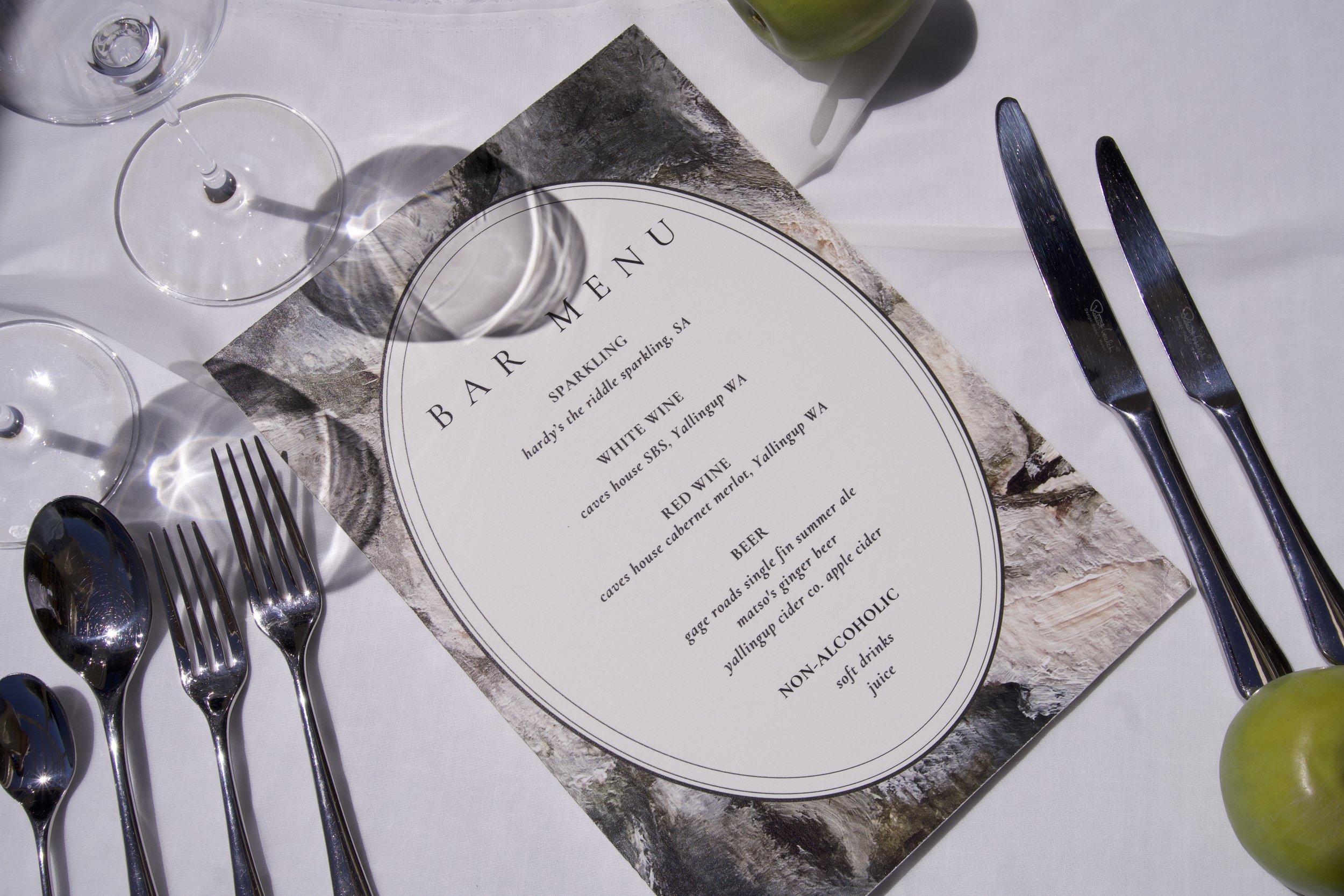 Table setting with a menu, wine glasses, forks, knives, and a green apple.