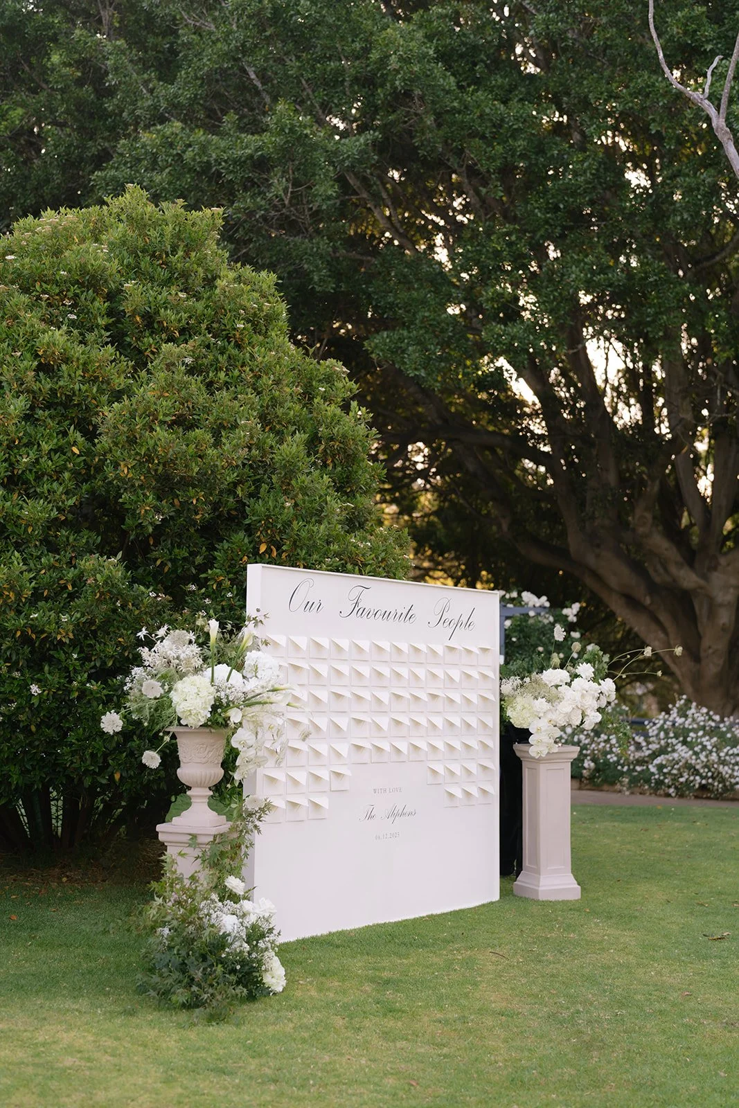Wedding seating chart display with white flowers on pedestals and lush greenery background.