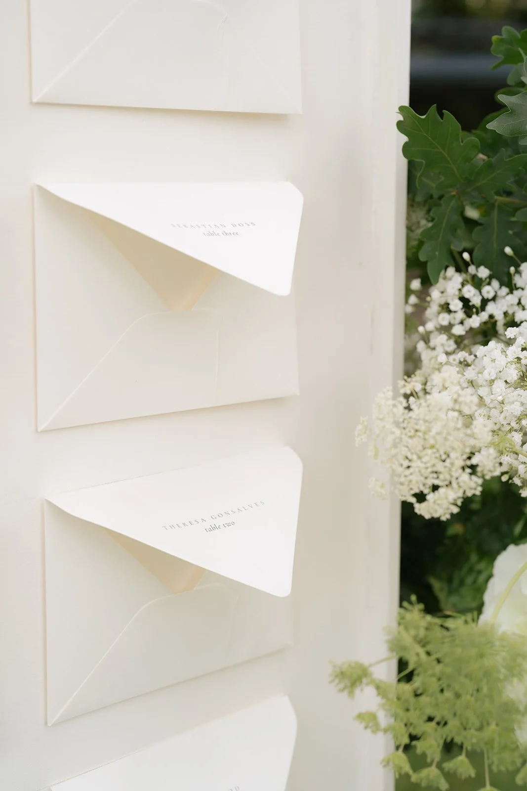 Three white envelopes with seating assignment cards inserted, attached to a white board, with green foliage and white flowers in the background.