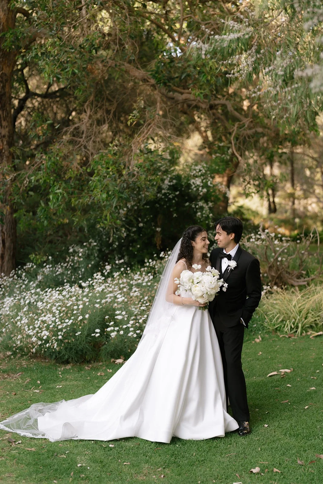 A bride and groom standing close together outdoors, smiling at each other. The bride is wearing a white wedding gown with a long train and carrying a bouquet of white flowers. The groom is in a black tuxedo with a white shirt and black bow tie. They are surrounded by greenery and small white flowers, with a large tree in the background.