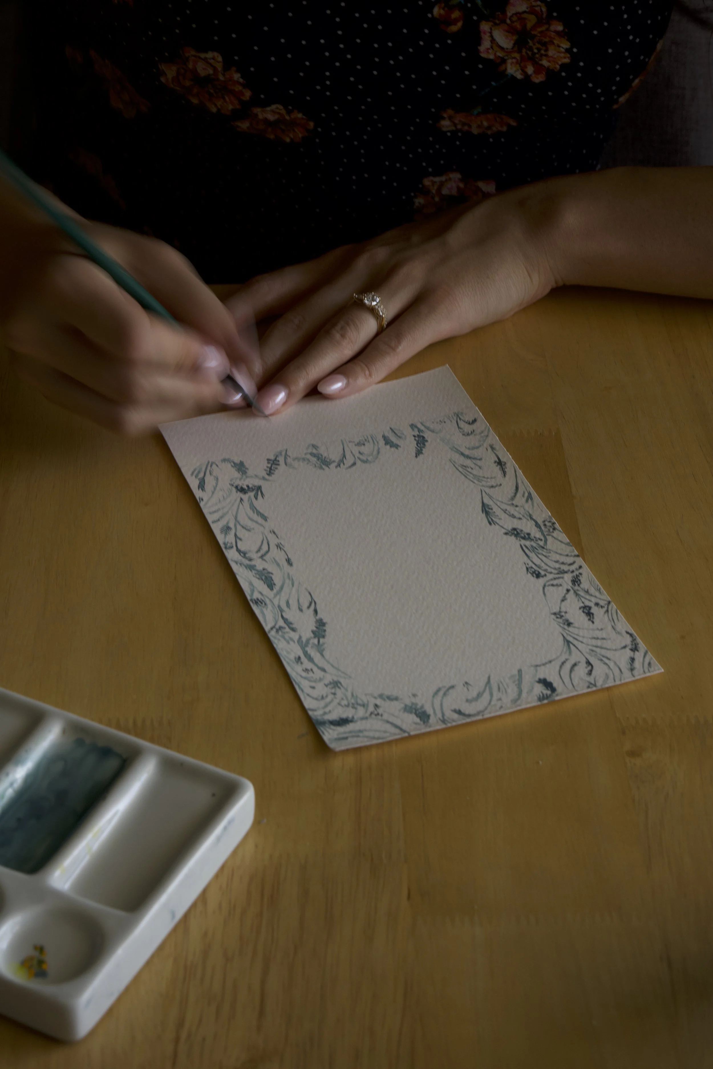 Person writing on a decorative blank card with a pen, wearing a ring, on a wooden table with watercolor paints in the corner.