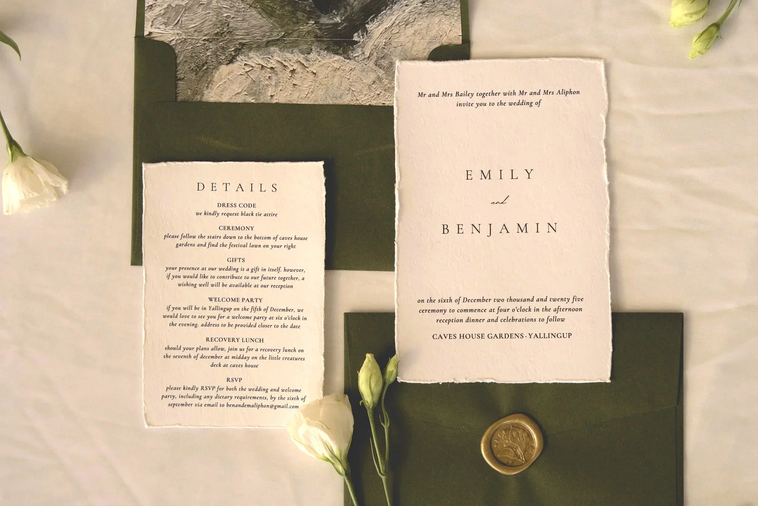Wedding invitation set including an outer envelope with a wax seal, a main invitation card, and a details card, all decorated with white flowers and placed on a white tablecloth.