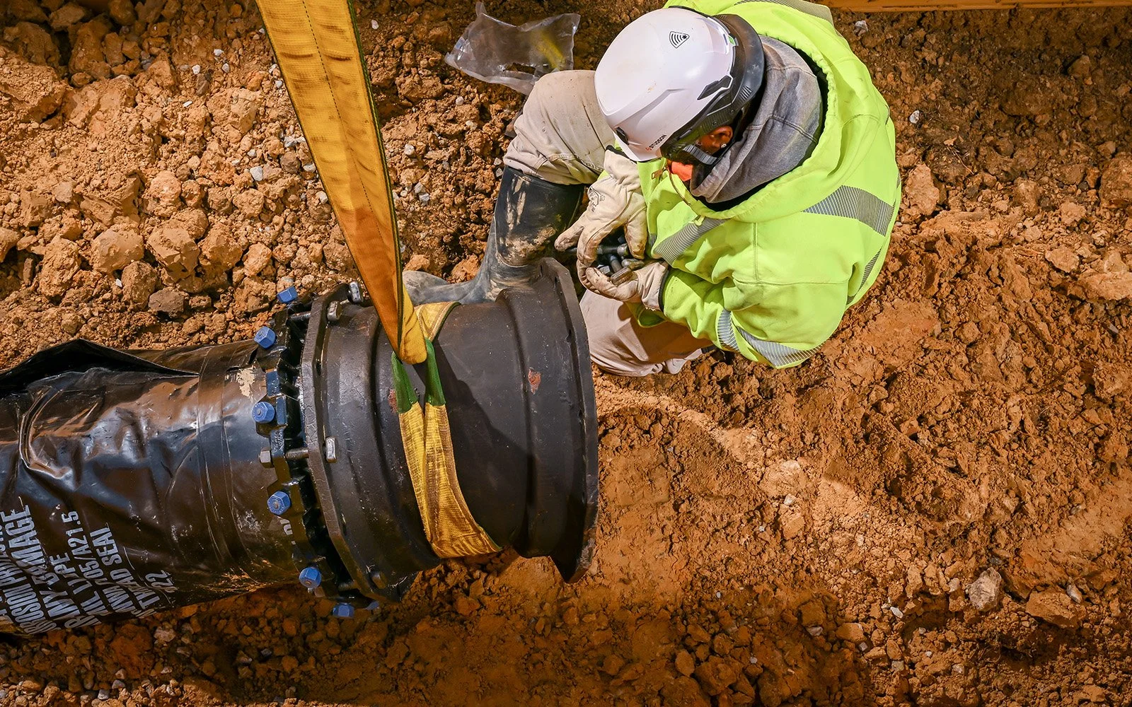 A construction worker in a high-visibility jacket and helmet is installing a large pipe or underground utility component at a construction site with dirt ground.
