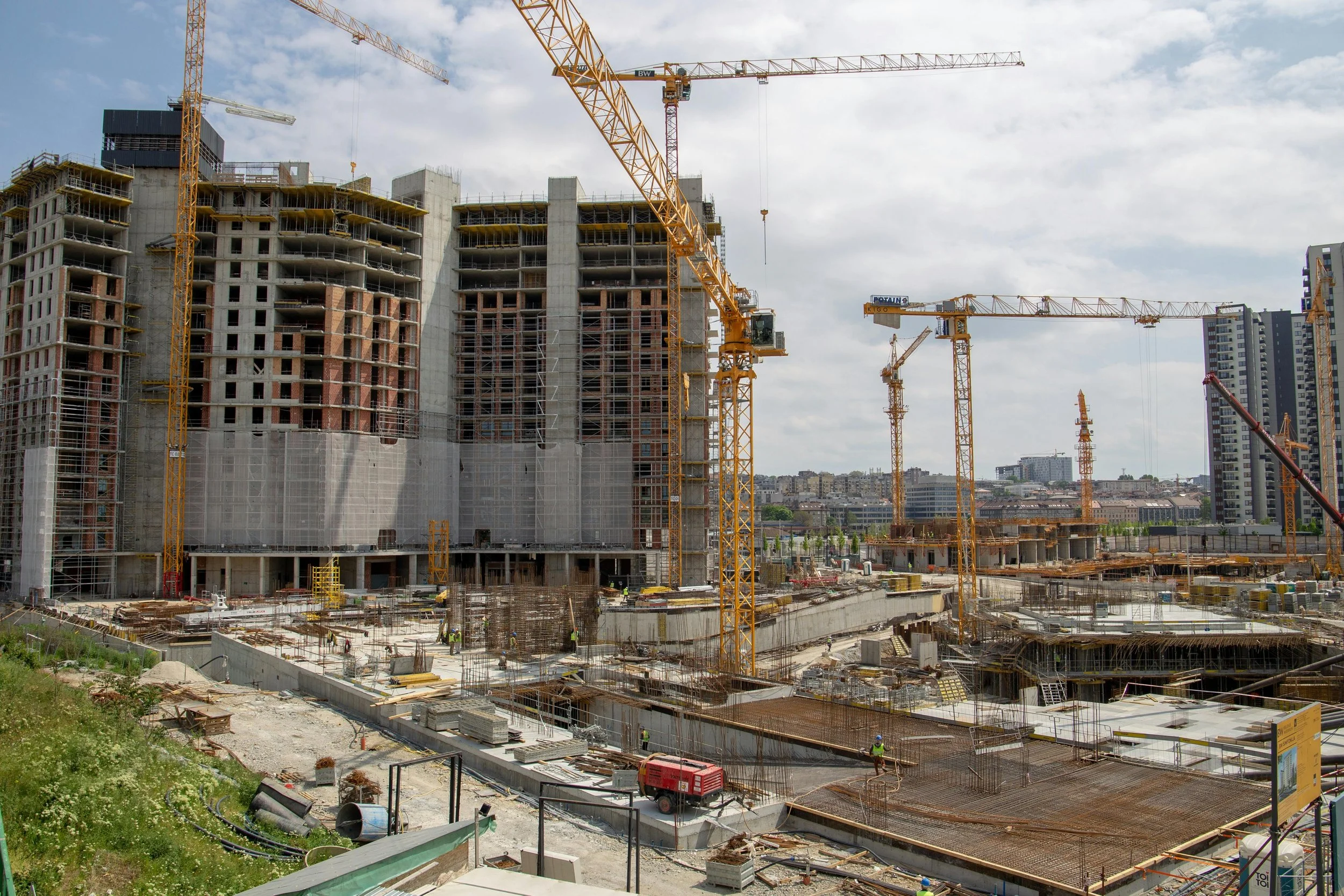 Construction site with multiple high-rise buildings under construction and cranes working on the site, with a cityscape in the background.