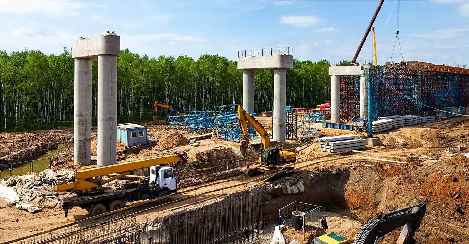 Construction site with large concrete pillars, cranes, and construction machinery, with trees in the background.