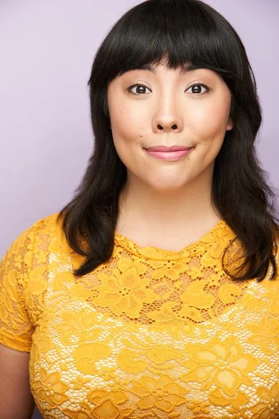 Headshot of Teresa Attridge, a woman with straight black hair and bangs, wearing a yellow lace top, smiling softly in front of a light purple background.