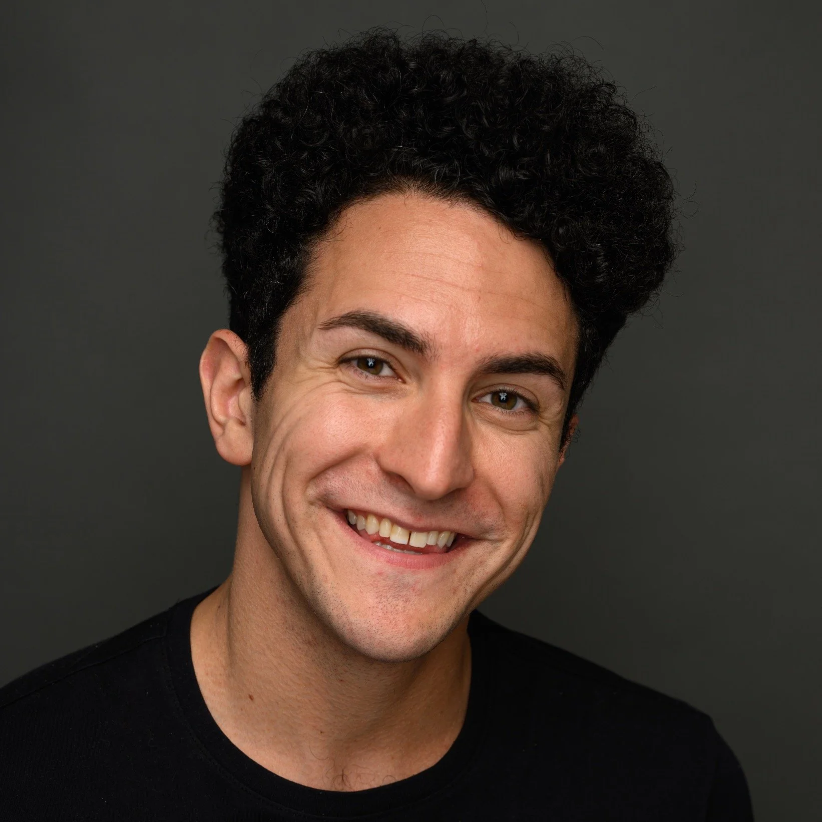 Headshot of Zach Faust, a smiling man with curly black hair wearing a black shirt against a dark background.
