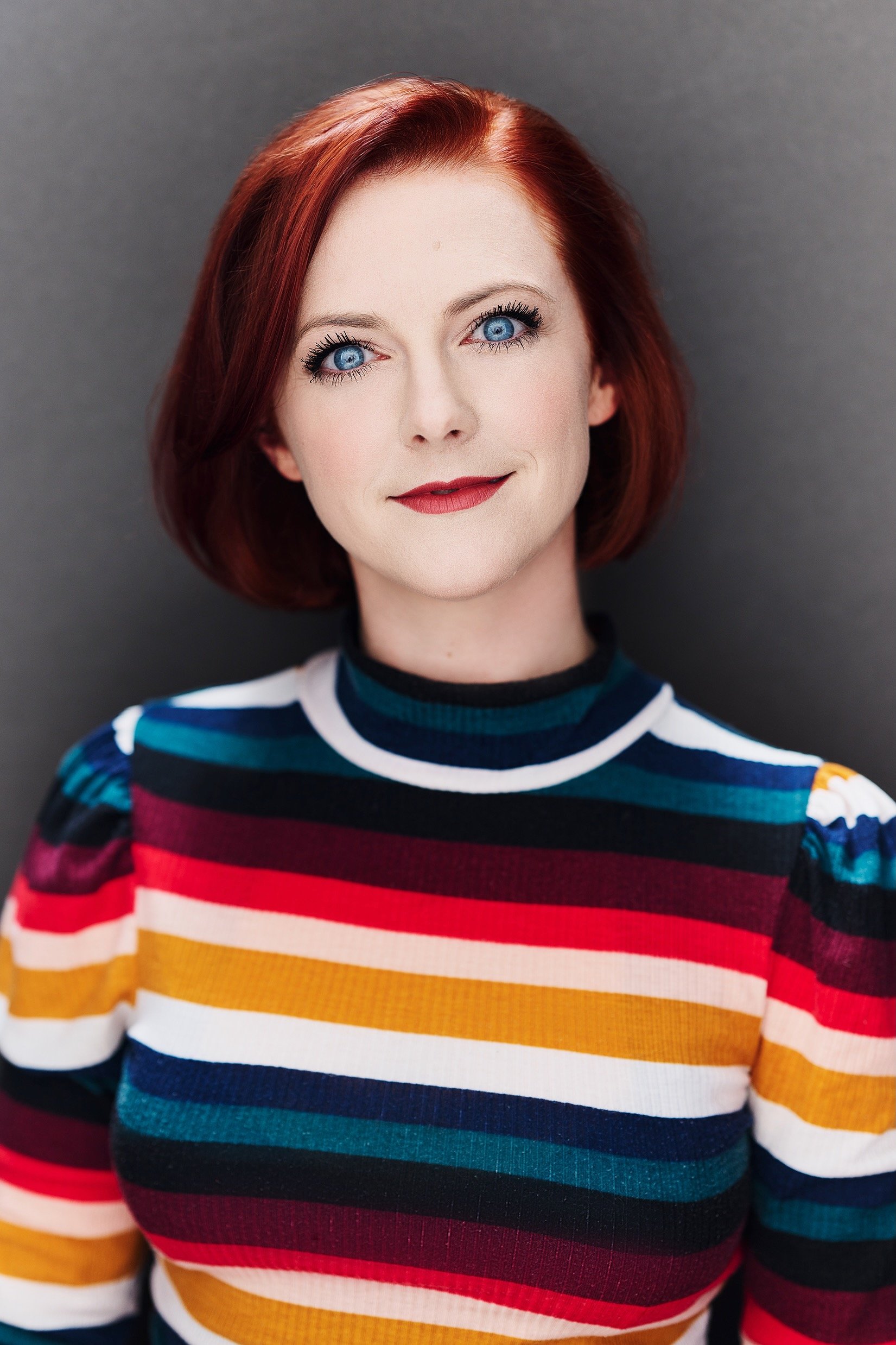 Headshot of Katie Emerson, a woman with red hair and blue eyes wearing a colorful striped sweater against a gray background.