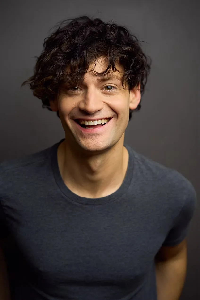 Headshot of Harrison Bryan, a man with curly dark hair, wearing a dark gray t-shirt, standing against a dark background.