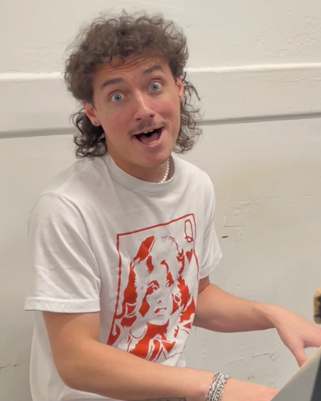 A young man with curly hair and a mustache, wearing a white T-shirt with red artwork, is sitting and looking excited while playing a keyboard. He is wearing a pearl necklace and bracelet, and the background is plain white.