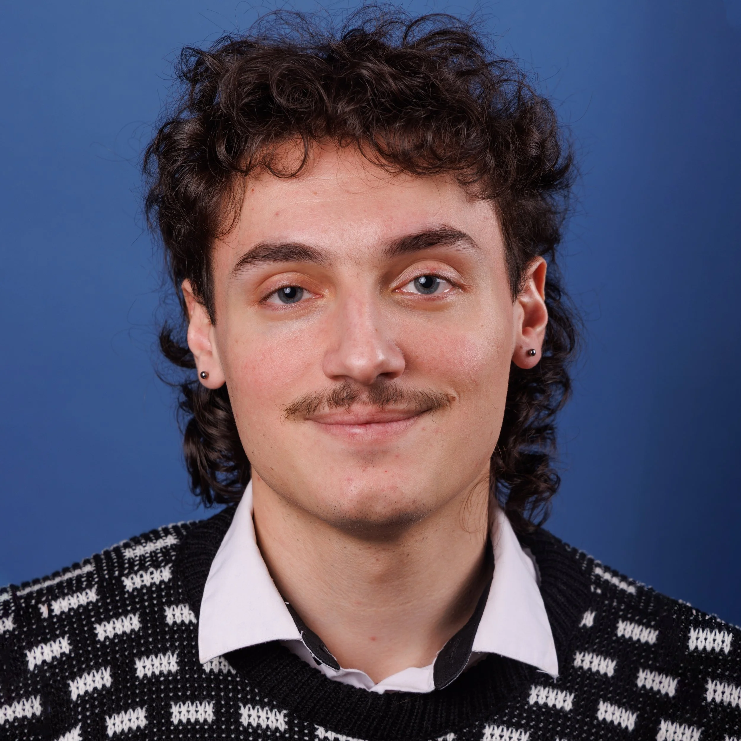 Justino Tesoro, a a young man with curly hair, wearing a white shirt and a black-and-white patterned sweater, against a blue background.
