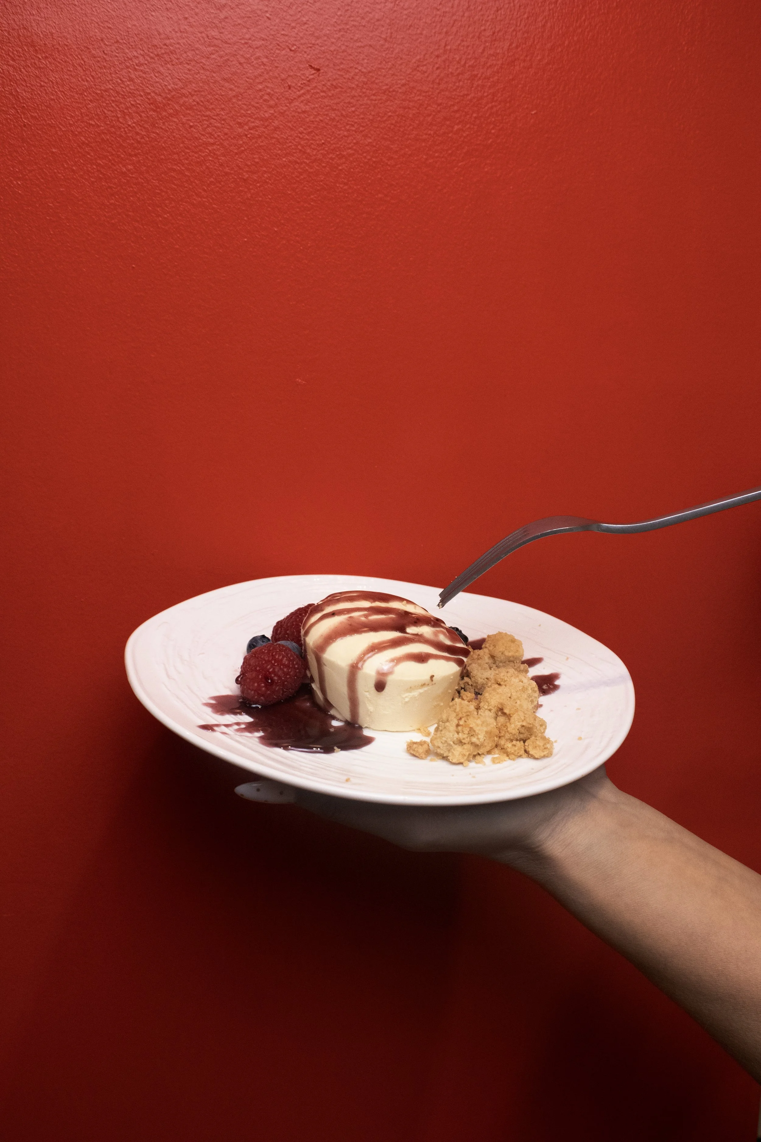 A white plate with a scoop of vanilla ice cream drizzled with chocolate syrup, fresh berries, and a piece of crumbly cookie or cake, held by a person's hand against a red background.
