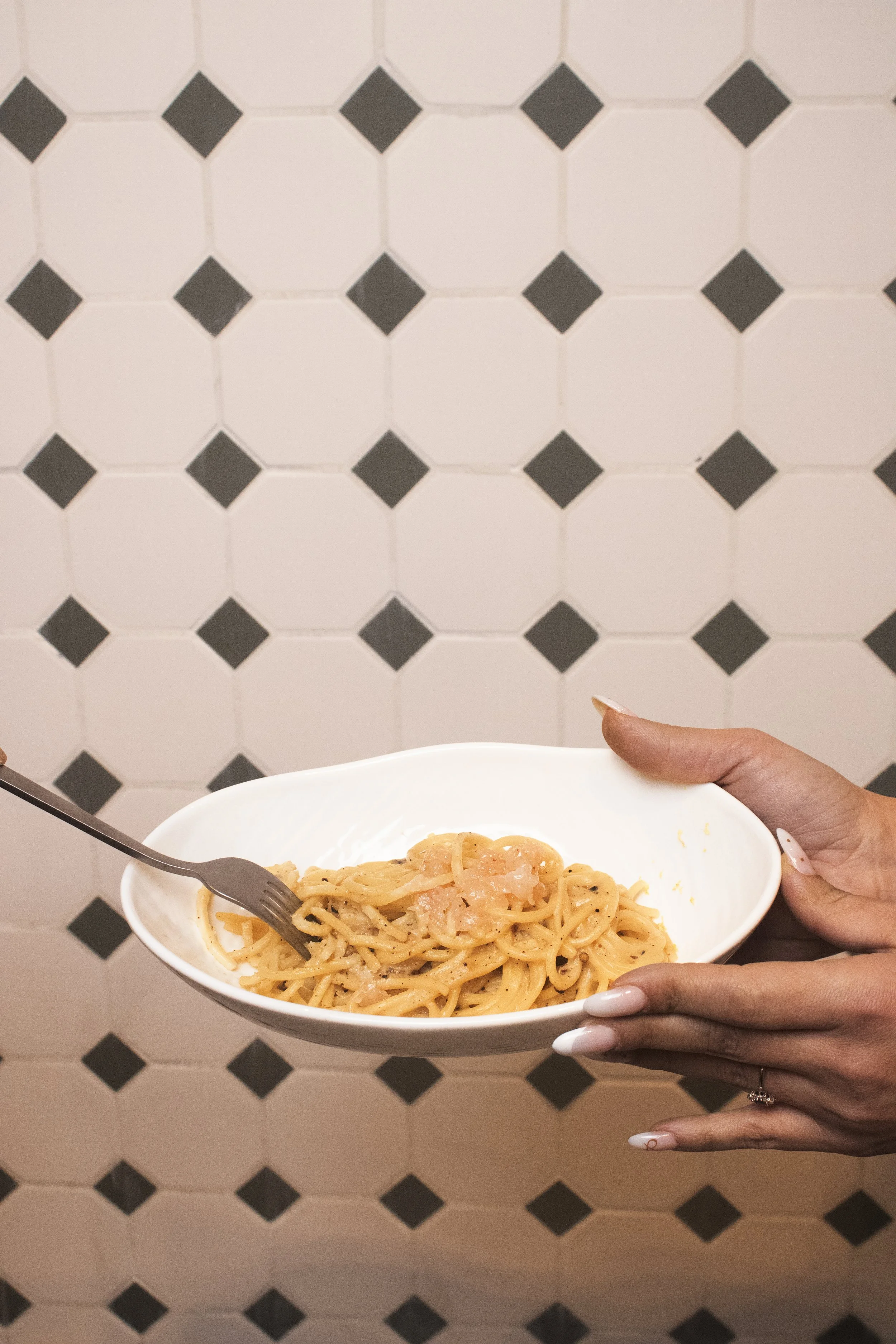 Person holding a white bowl of pasta with a fork in front of a tiled wall.