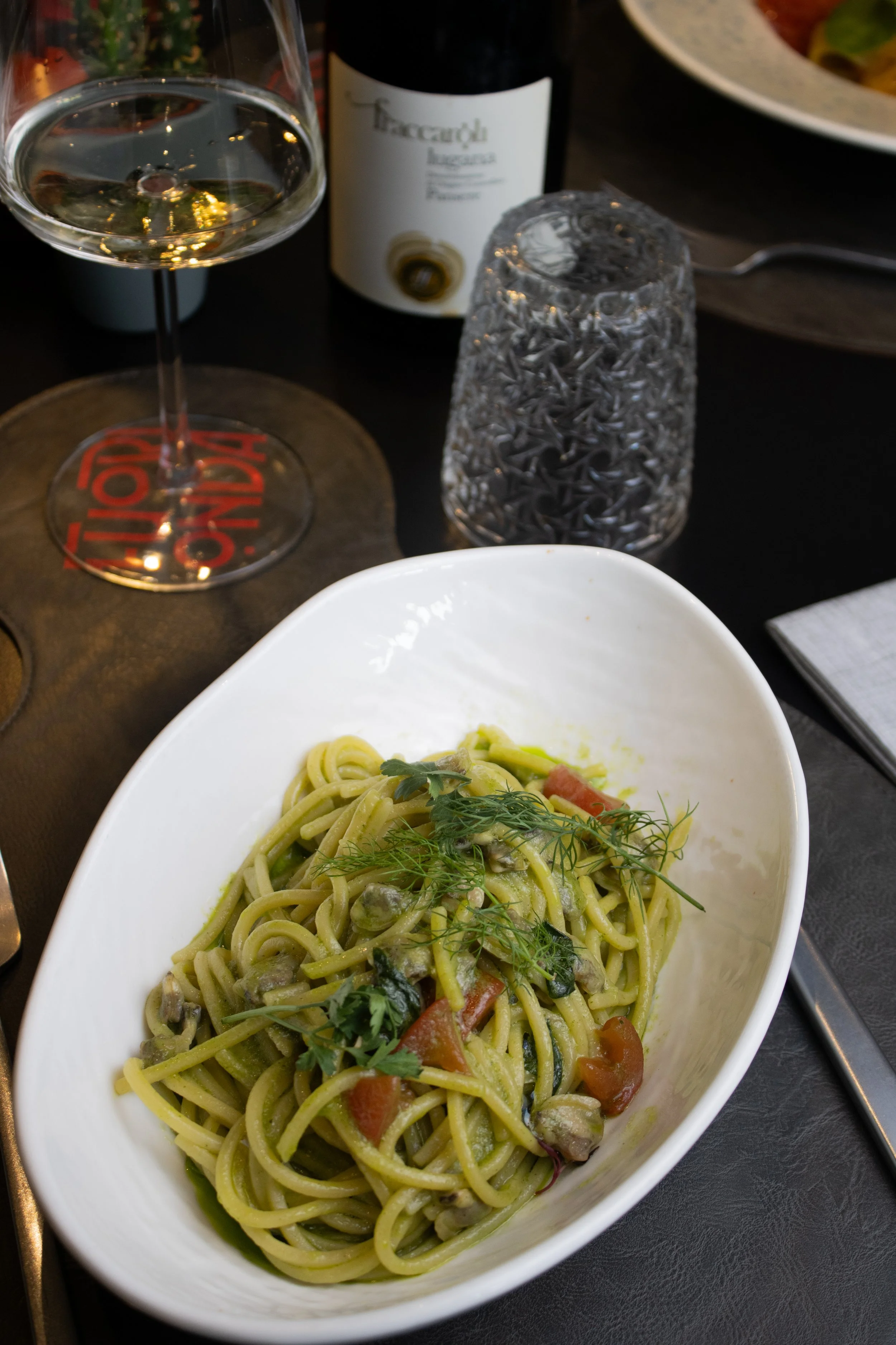 Una ciotola bianca di pasta con pomodorini, erbe aromatiche e verdure. Sullo sfondo si vedono un calice di vino bianco, una bottiglia di vino, un bicchiere trasparente lavorato e una padella nera su un tavolo nero.