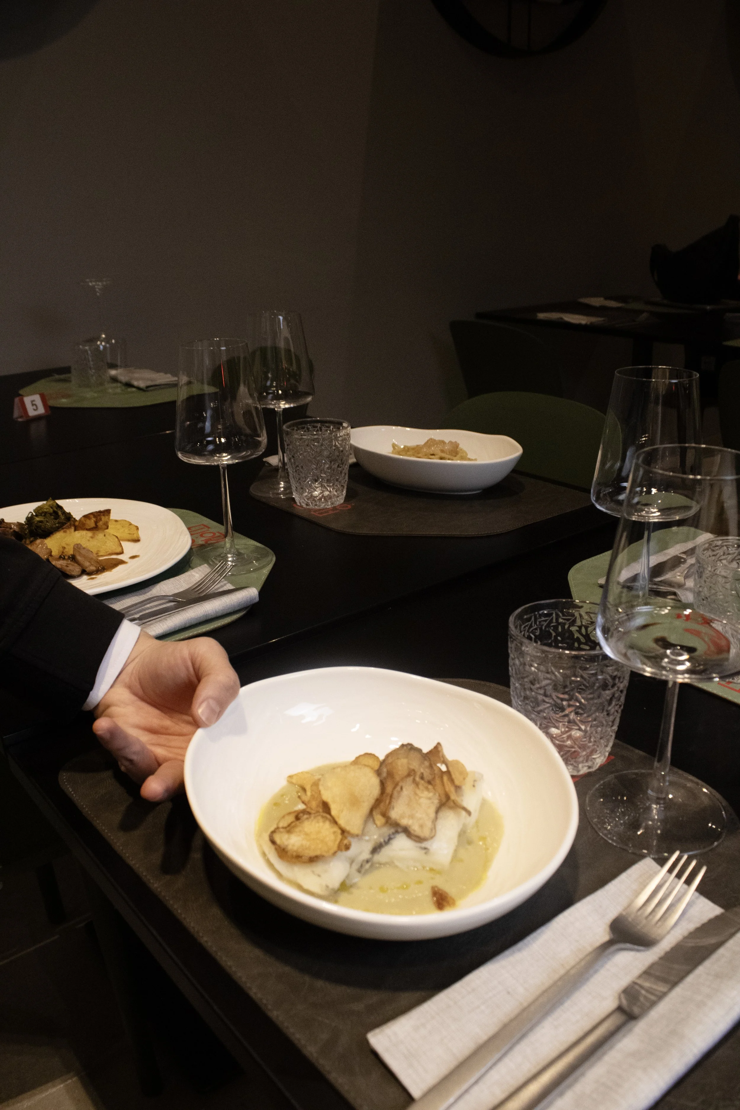 A person holding a white bowl with a piece of fish topped with potato chips, served in a cream sauce, on a dining table with empty glasses, utensils, and additional dishes in the background.