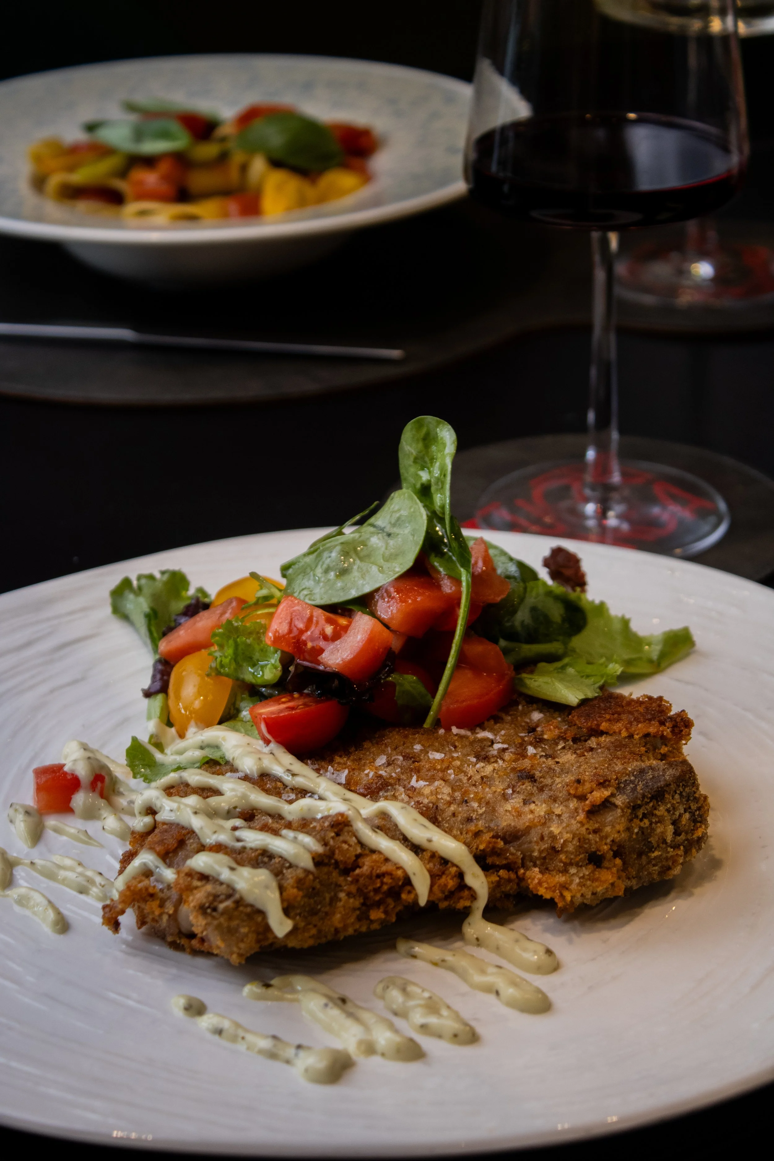 Piatto con filetto di pesce impanato, guarnito con pomodori a cubetti, verdure e condimento, accompagnato da un calice di vino rosso in un ambiente dalla luce soffusa.