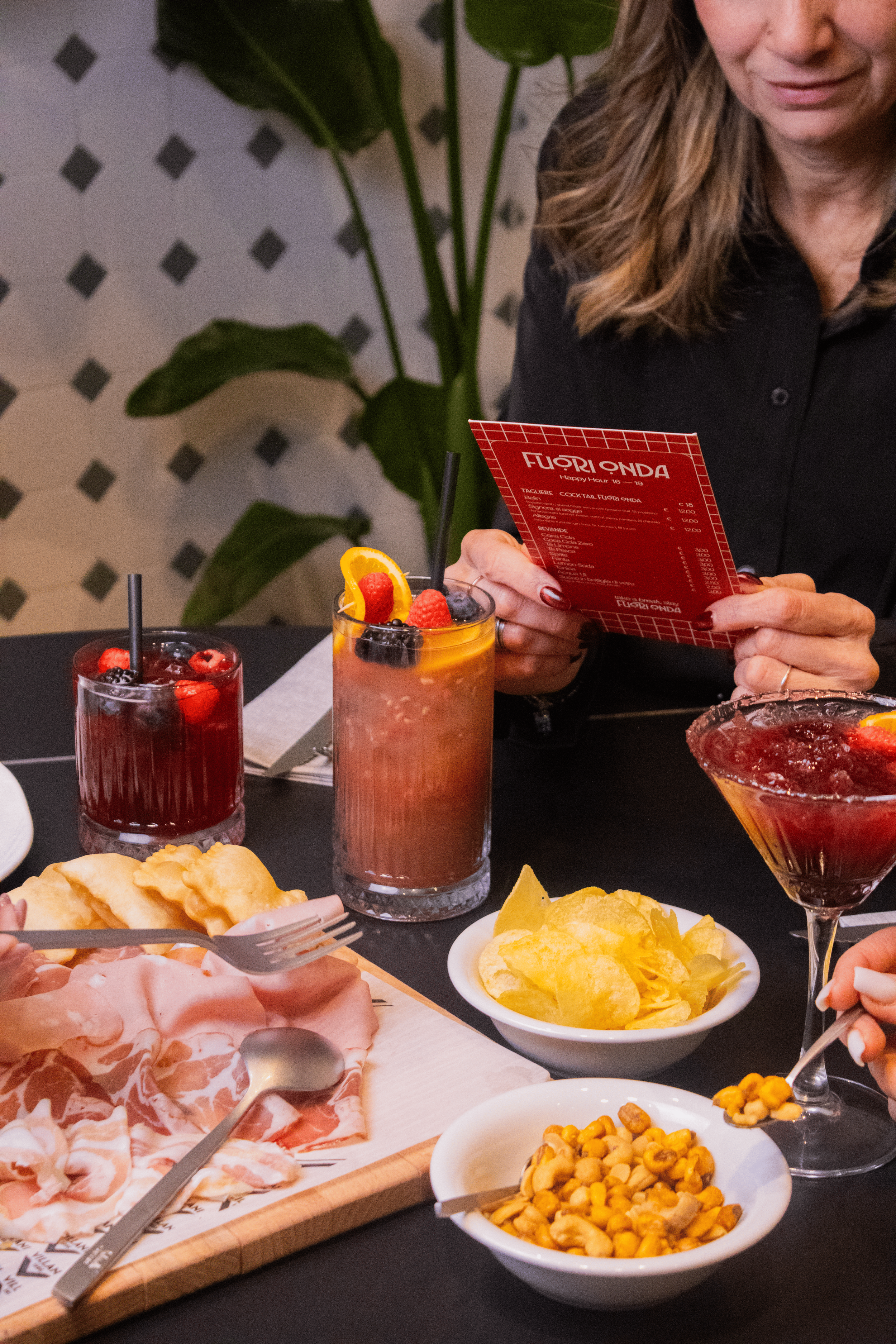 A woman at a table with various dishes and drinks, holding a menu and smiling. The table has chips, cured meats, fruit cocktails, and a bowl of nuts.