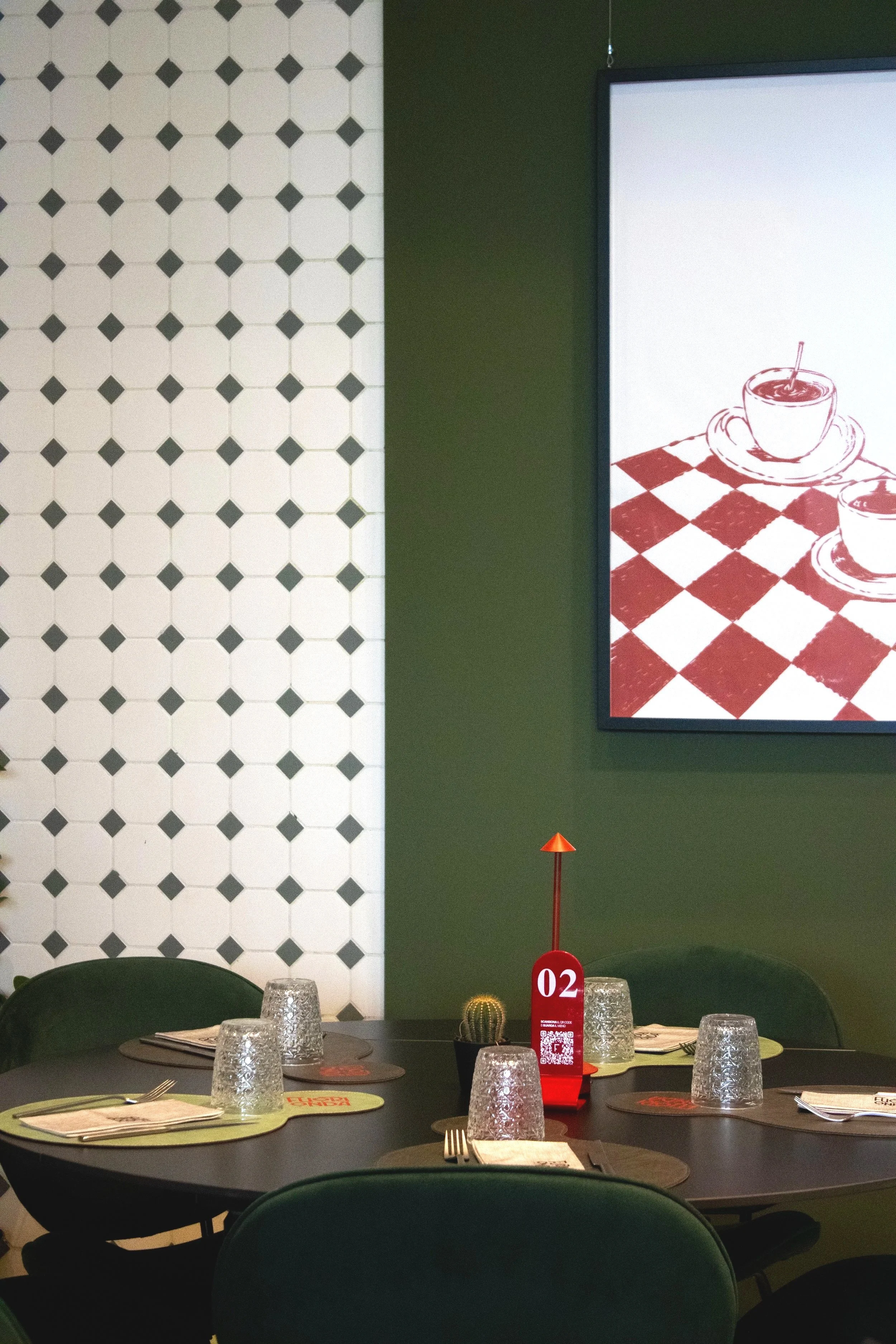 Interno di un ristorante con un tavolo rotondo apparecchiato con bicchieri, piatti e posate. Al centro si trovano una piccola pianta di cactus e un segnaposto rosso. La parete sullo sfondo ha una sezione verde con un quadro raffigurante due tazze su