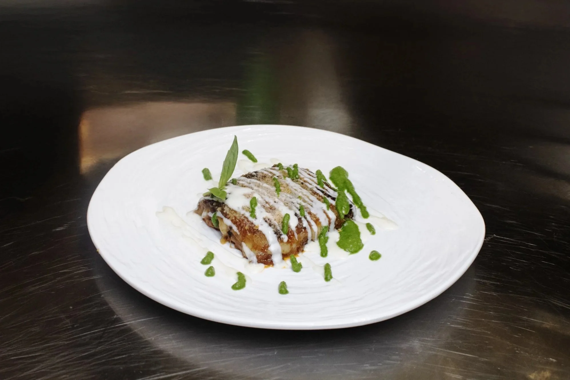 A plated dessert with a pastry topped with white icing, green sauce, and garnished with herbs on a white plate against a dark background.