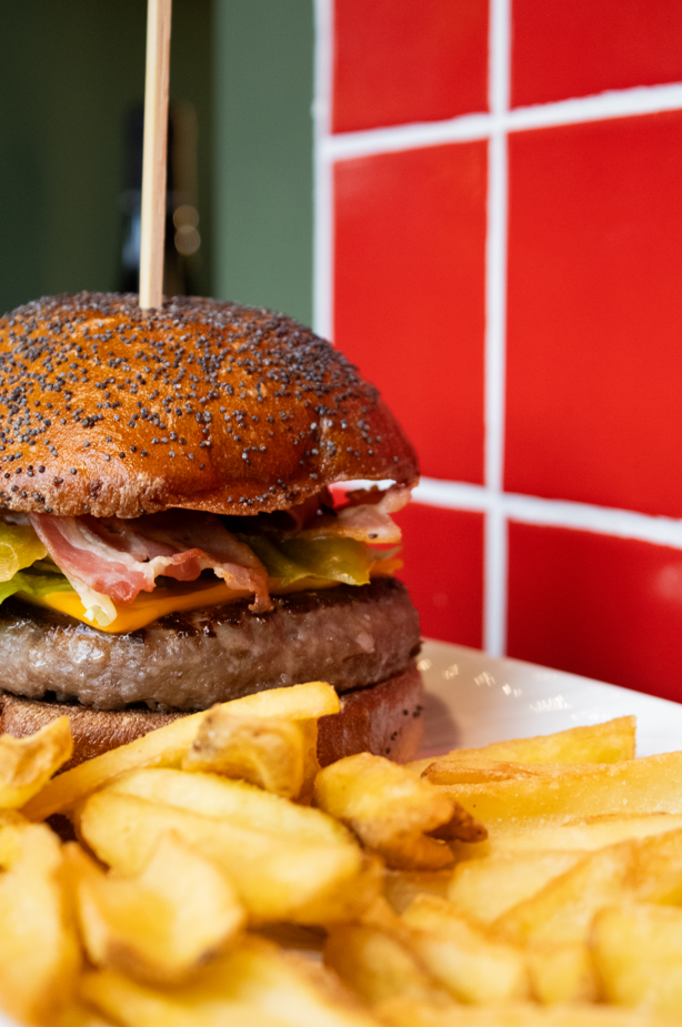 Primo piano di un cheeseburger con bacon, cetriolini e lattuga in un panino ai semi di papavero, servito con un contorno di patatine fritte su un piatto bianco. Sullo sfondo si vede una parete rivestita di piastrelle rosse.