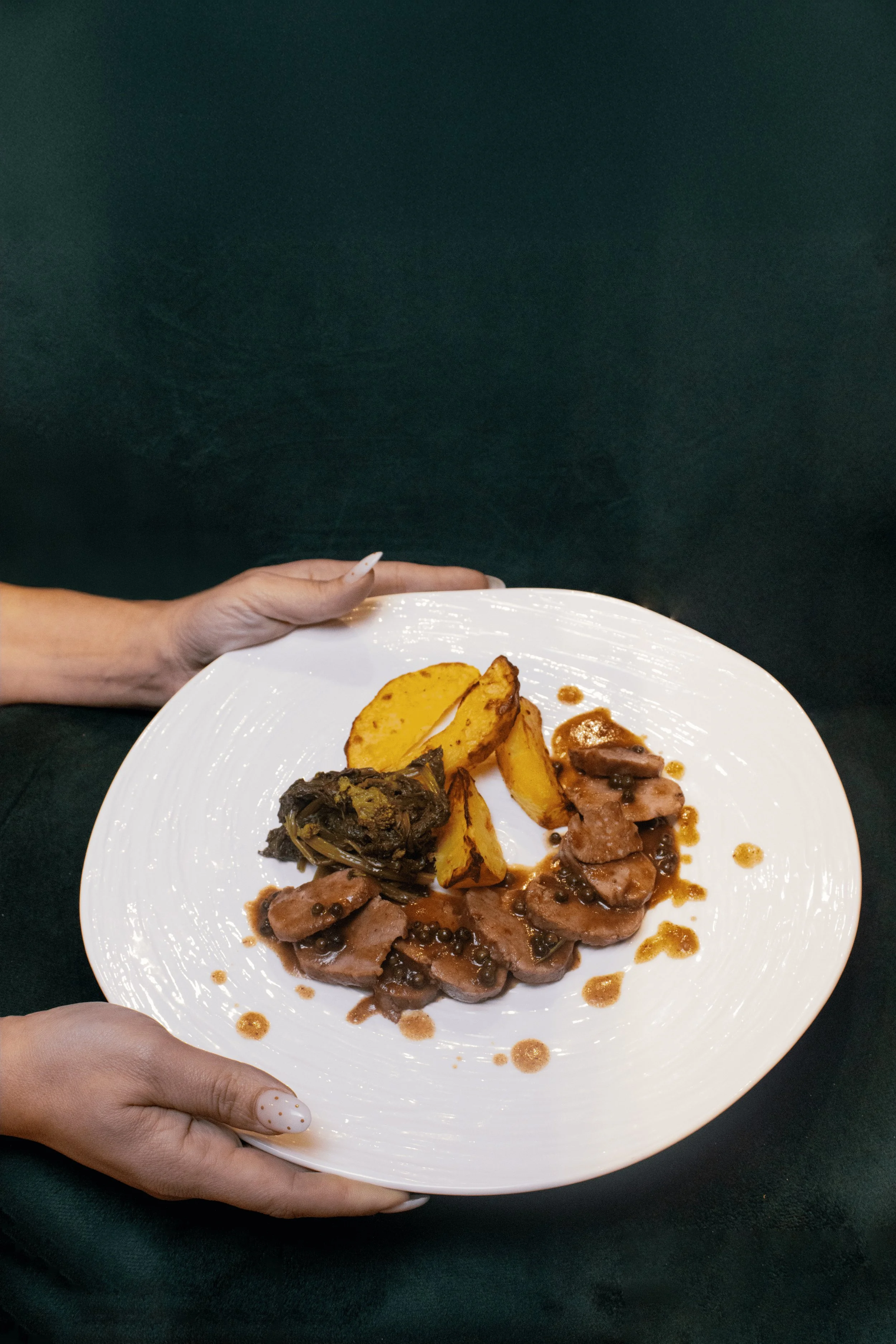 Hand holding a white plate with roasted potatoes, cooked meat with gravy, and a braised vegetable on a dark background.