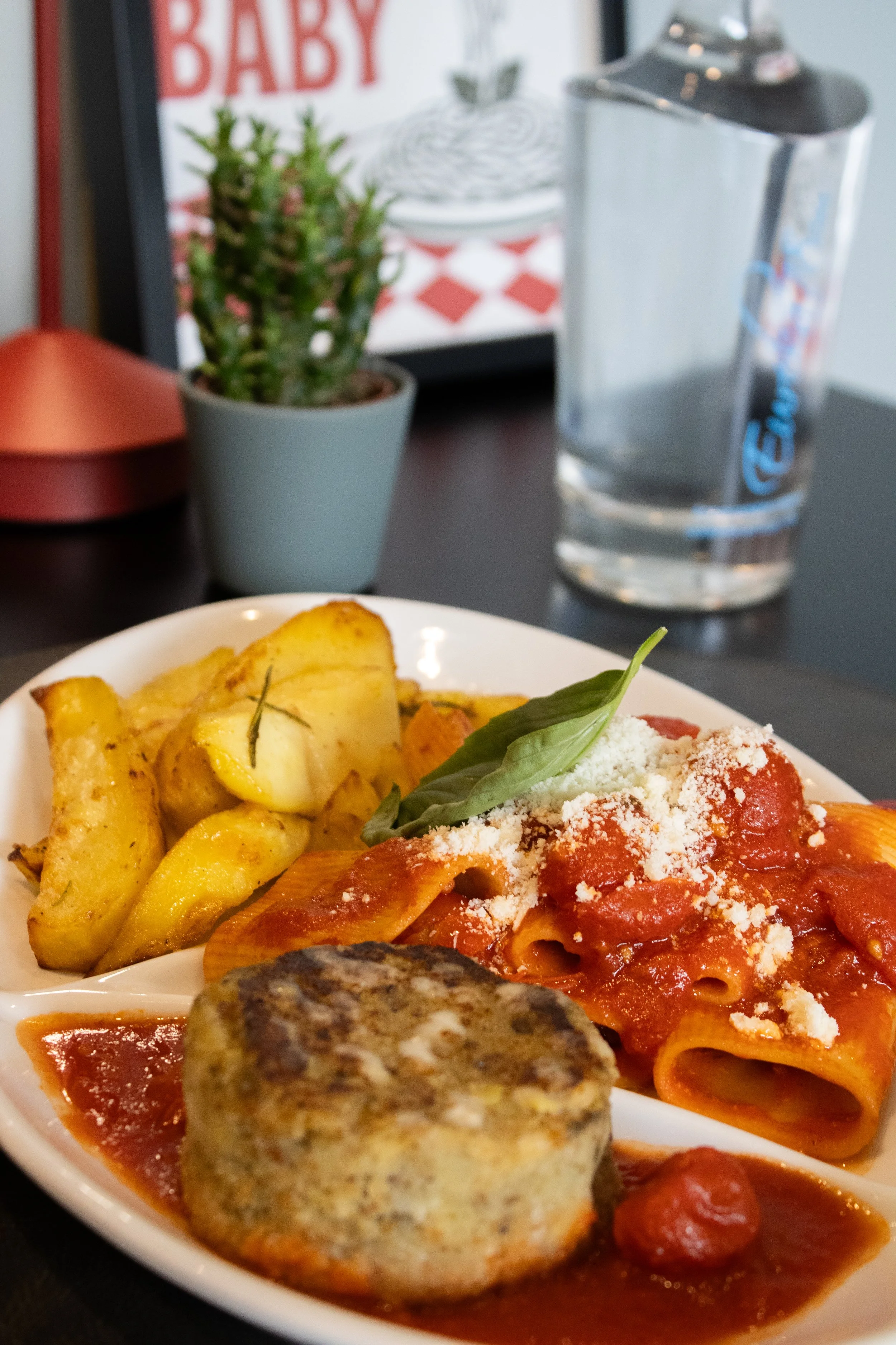 Un piatto di cucina italiana con pasta al pomodoro, formaggio grattugiato e basilico, accompagnata da patate arrosto e polpette. Sullo sfondo si vedono un bicchiere d’acqua, una pianta in vaso, una cornice con la scritta “BABY” e un contenitore decor