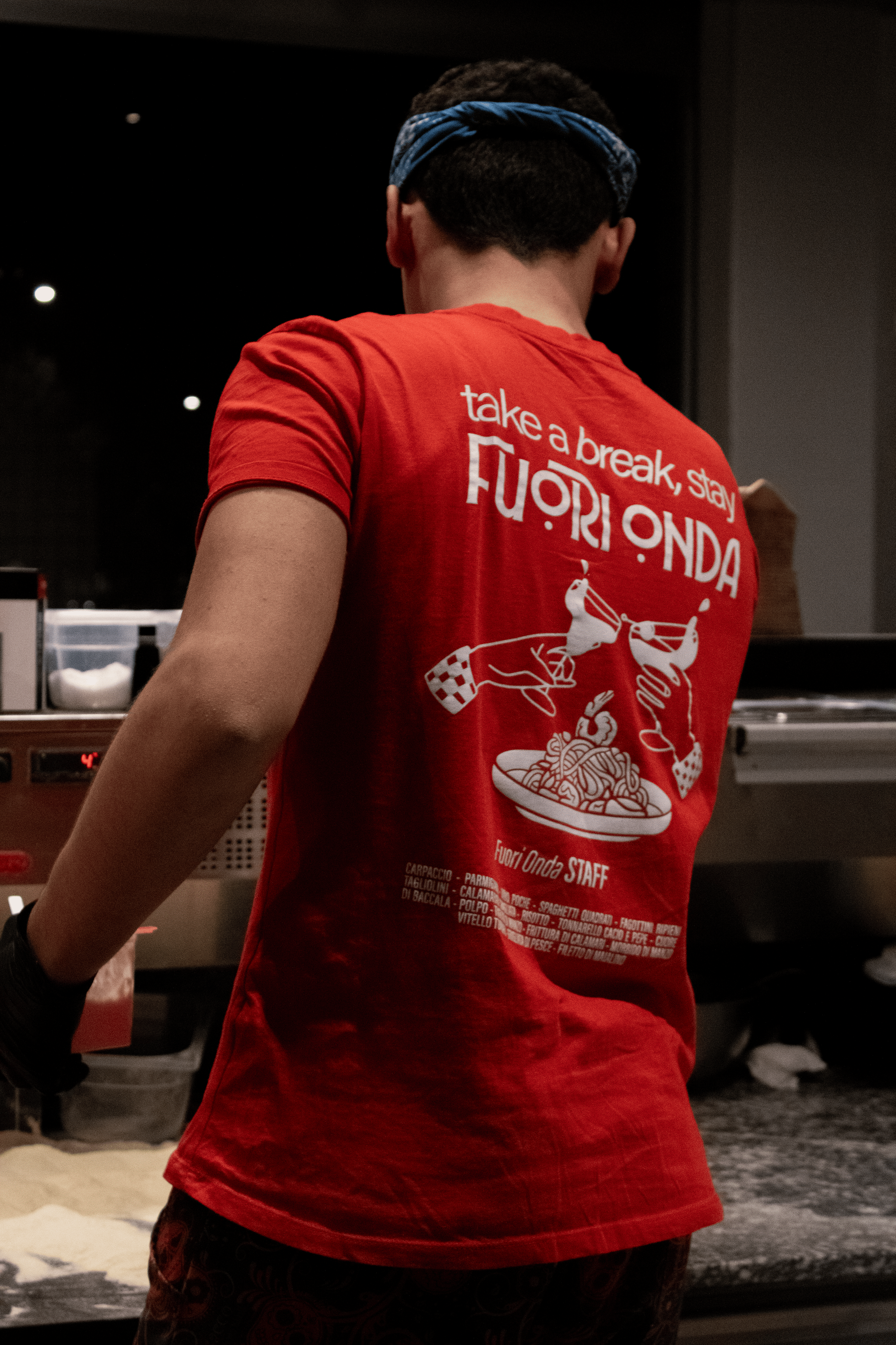 Back view of a person in a red T-shirt with white graphic and text, wearing a blue bandana, cooking or working at a food stall at night.