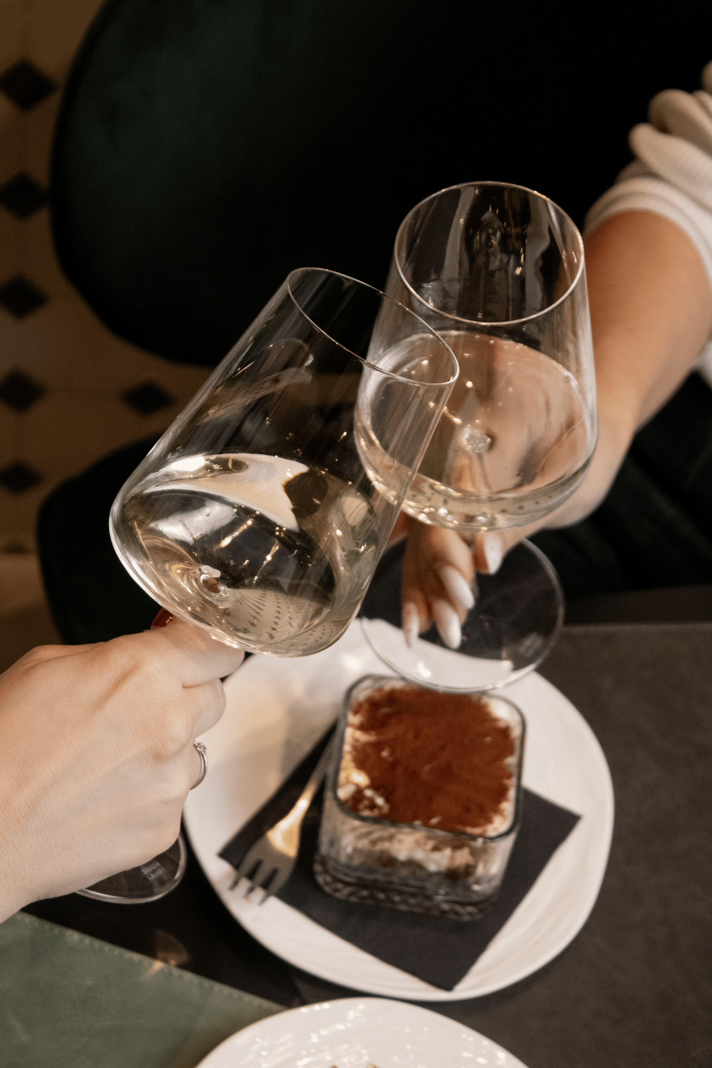 Two women clinking glasses of white wine at a dinner table with dessert and silverware.
