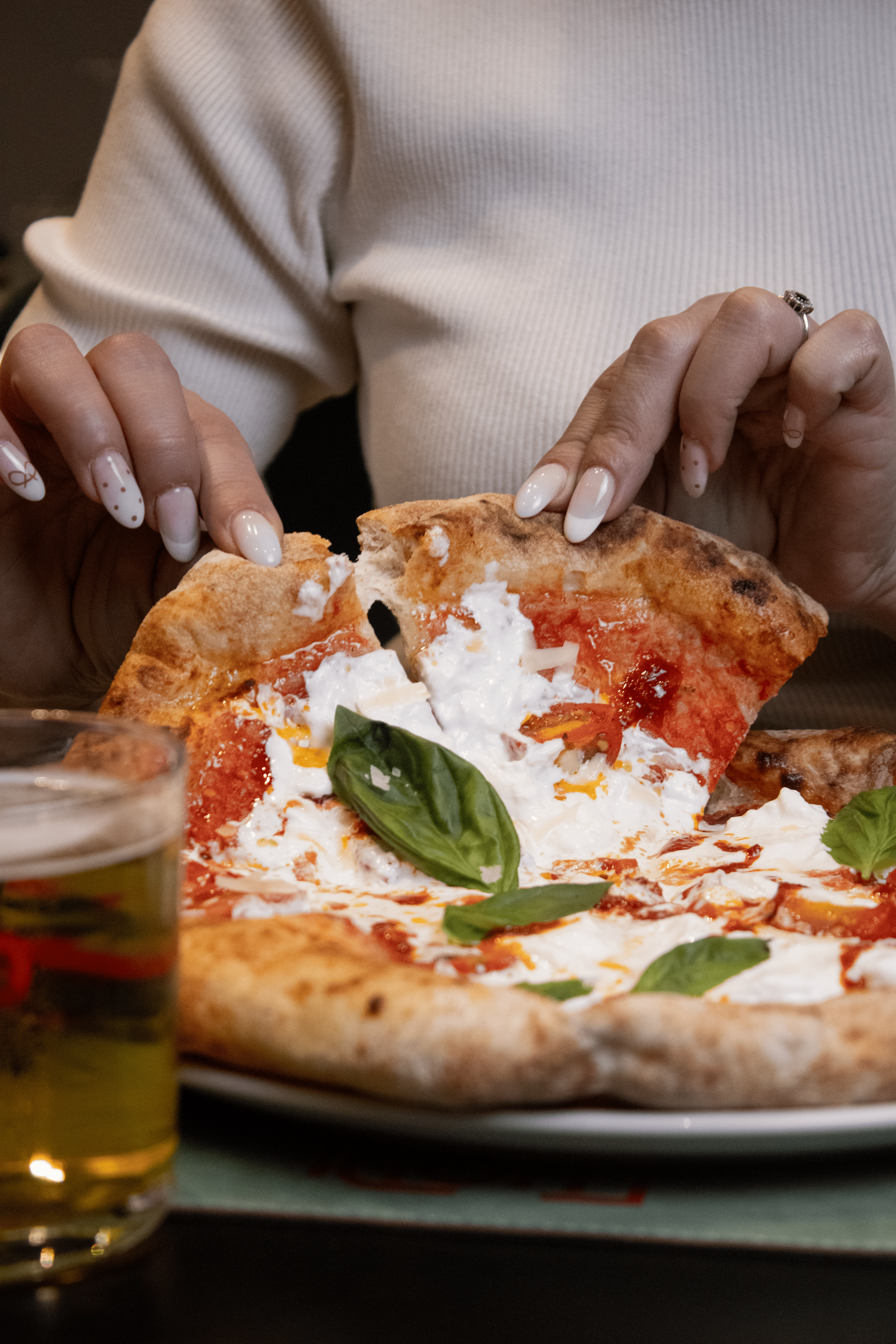 Person tearing a slice of pepperoni pizza with basil leaves on top, with a glass of beer nearby.