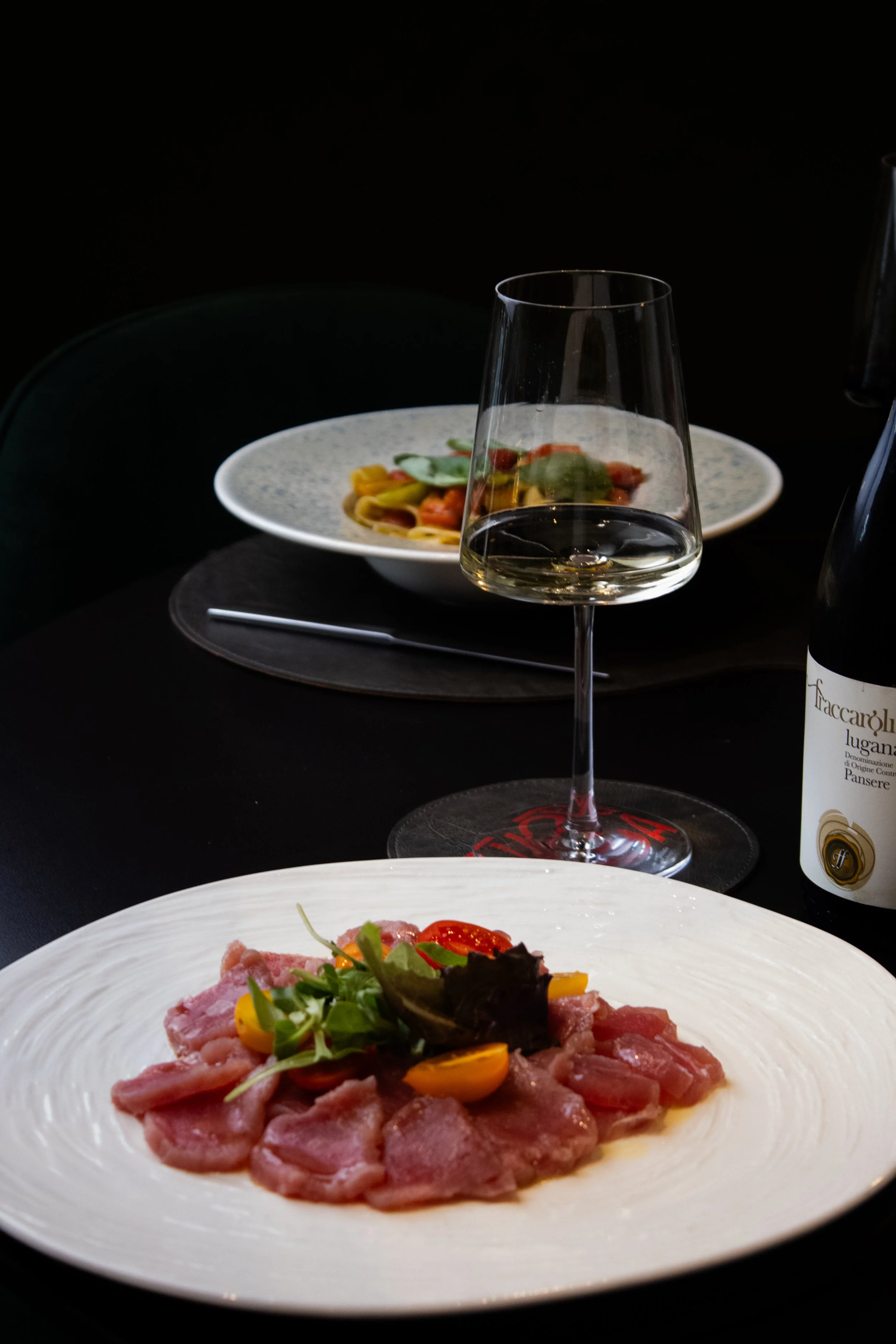 Un piatto bianco con carne cruda affettata, guarnita con verdure, pomodorini e fettine di peperone giallo. Sullo sfondo si vedono un bicchiere d’acqua e una bottiglia di vino su un tavolo scuro.