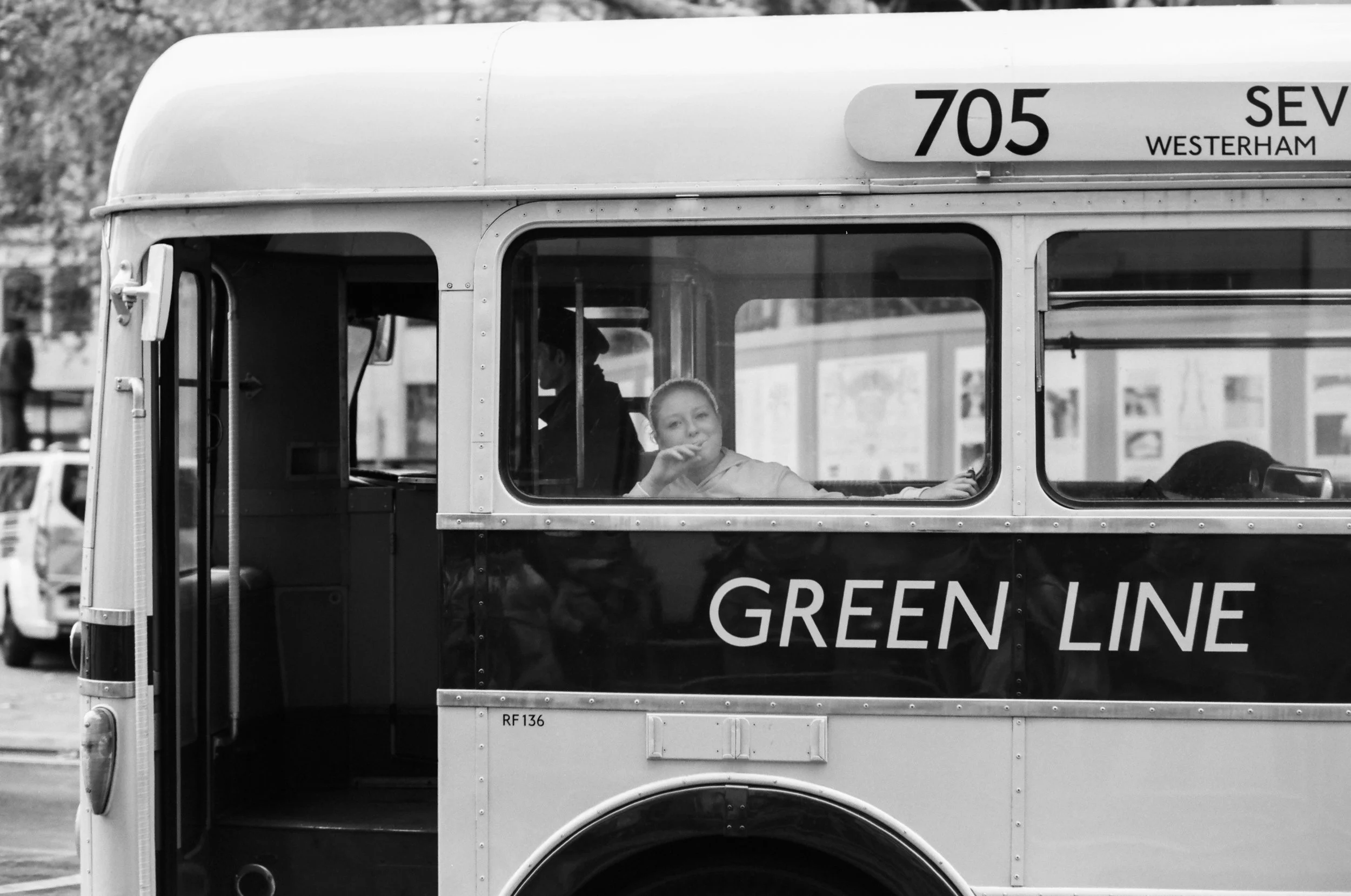 A black-and-white photo of a vintage double-decker bus labeled "Green Line" with a girl sitting by a window, looking outside and smiling, in an urban setting.