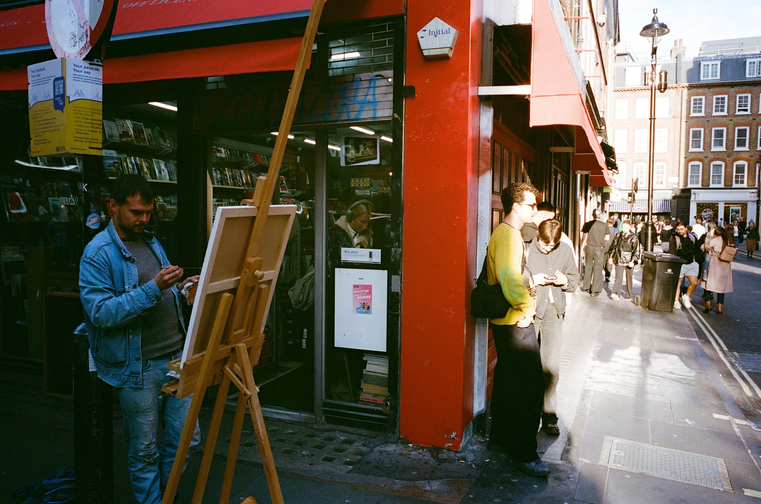People standing and socializing on a busy city street outside a shop with books and art supplies, with a man reading a book near an easel in front of a bookstore.