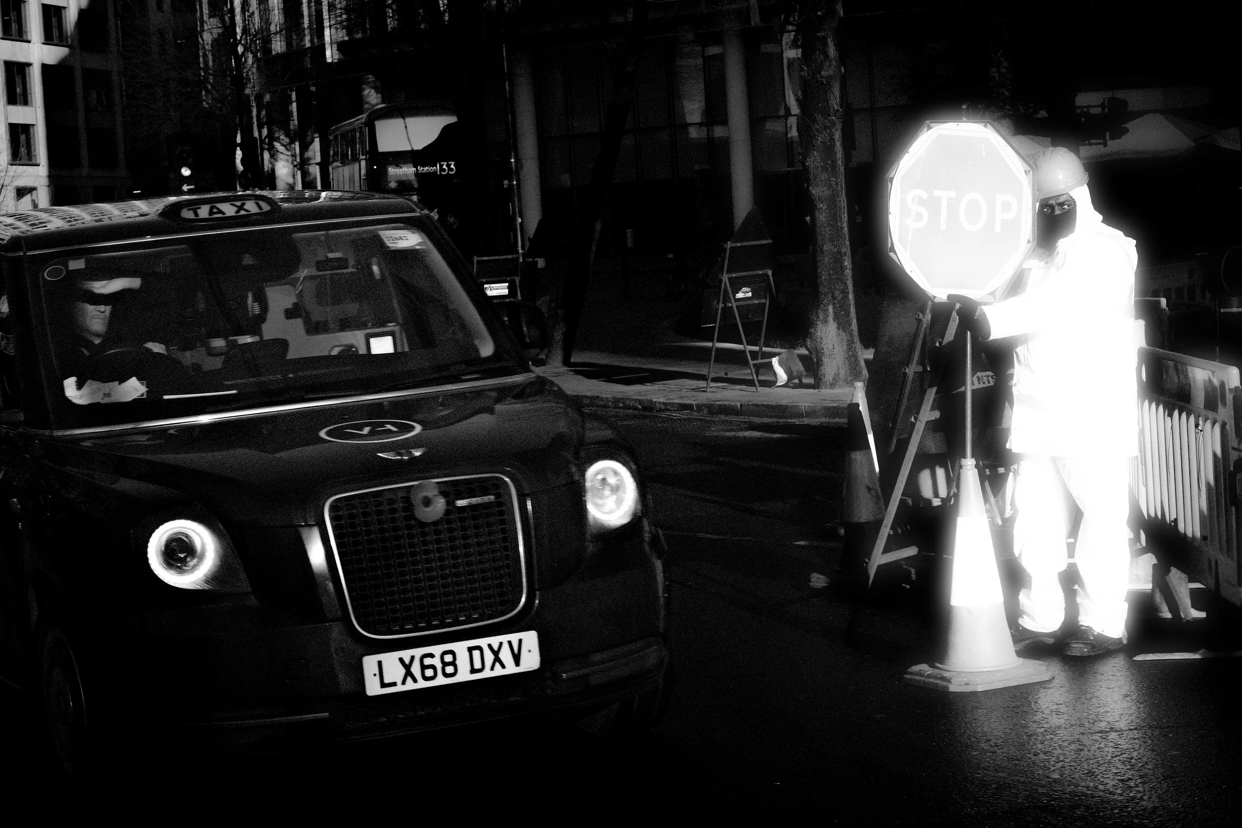 Nighttime scene with a black taxi cab and a worker wearing white protective clothing and a helmet, holding a reflective STOP sign. The worker is standing near orange cones and safety barriers, with trees and a building in the background.