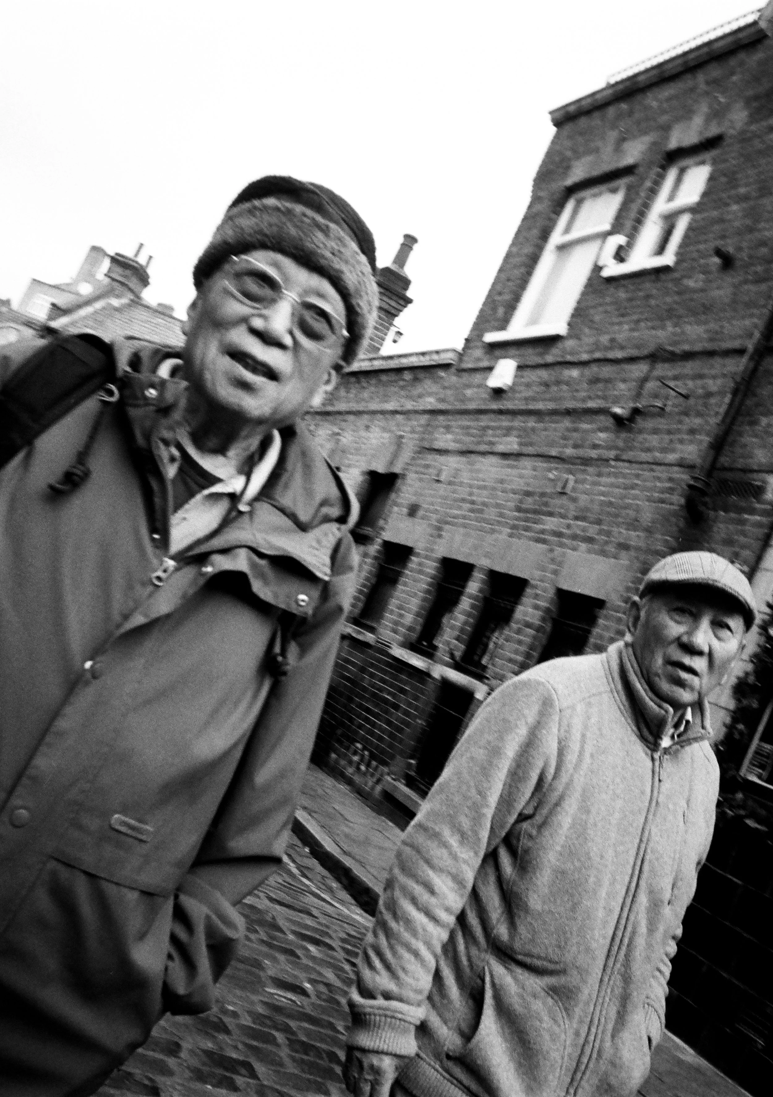 Two elderly men standing on a cobblestone street with brick buildings in the background, black and white photo.