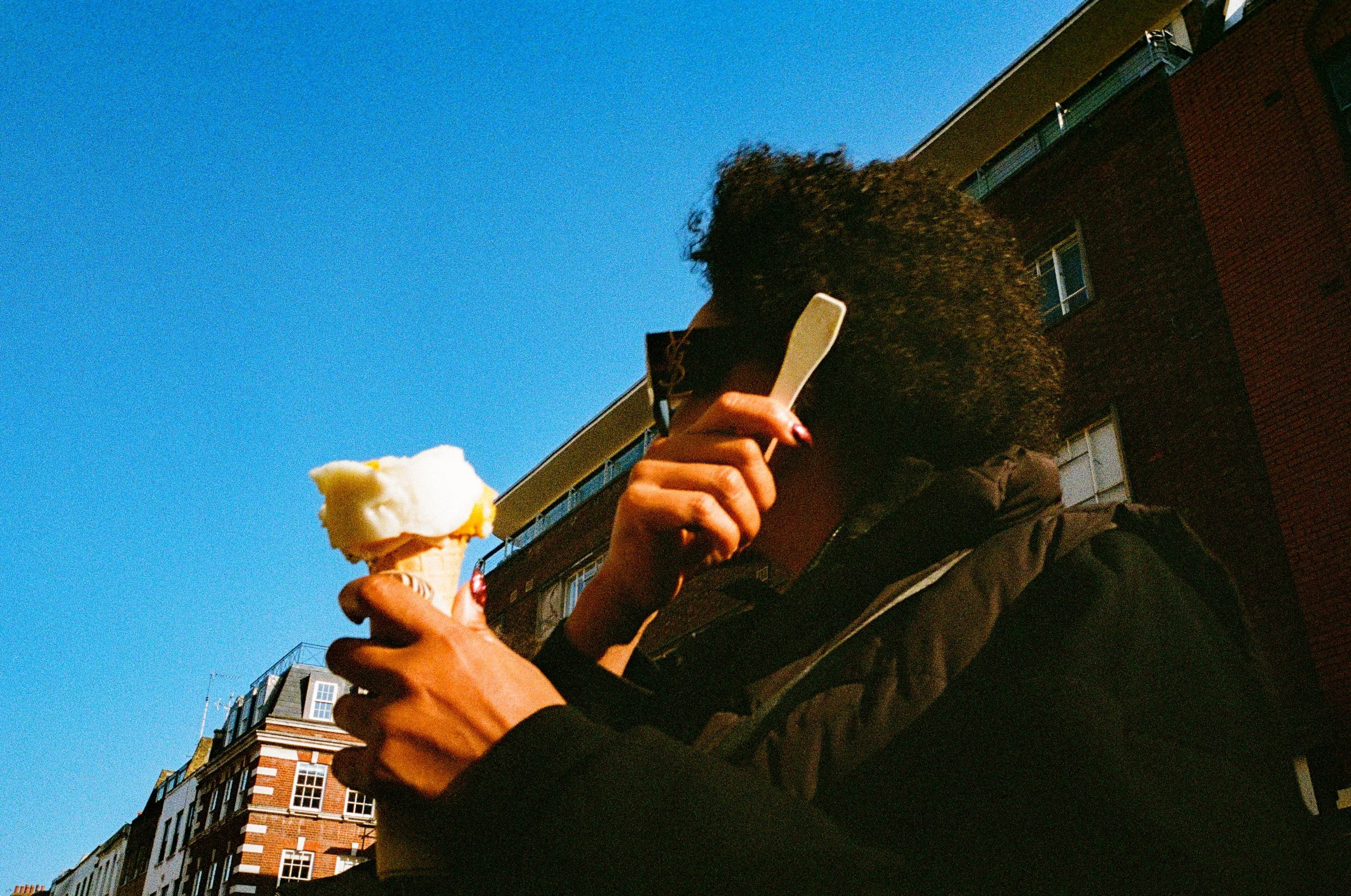 Person with curly hair wearing sunglasses and a dark jacket is eating ice cream cone and talking on a mobile phone on a city street with buildings and a clear blue sky in the background.