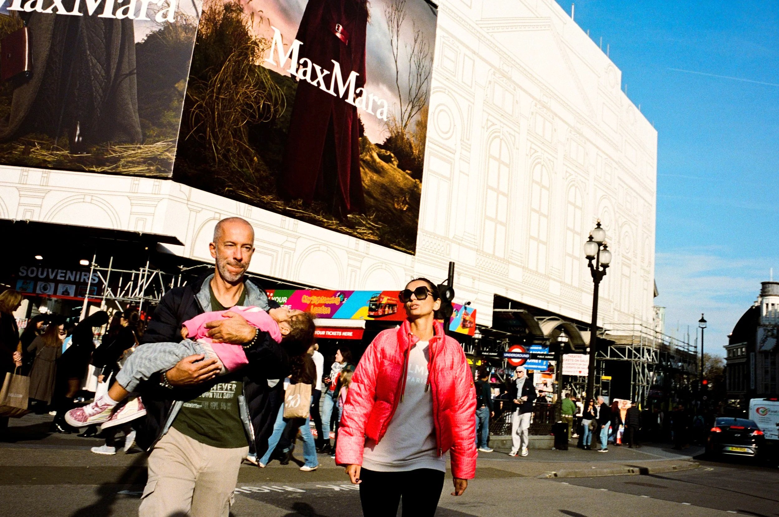 A man holding a sleeping child, a woman walking on a busy street in front of a large advertising billboard for MaxMara, with many pedestrians and city buildings in the background.