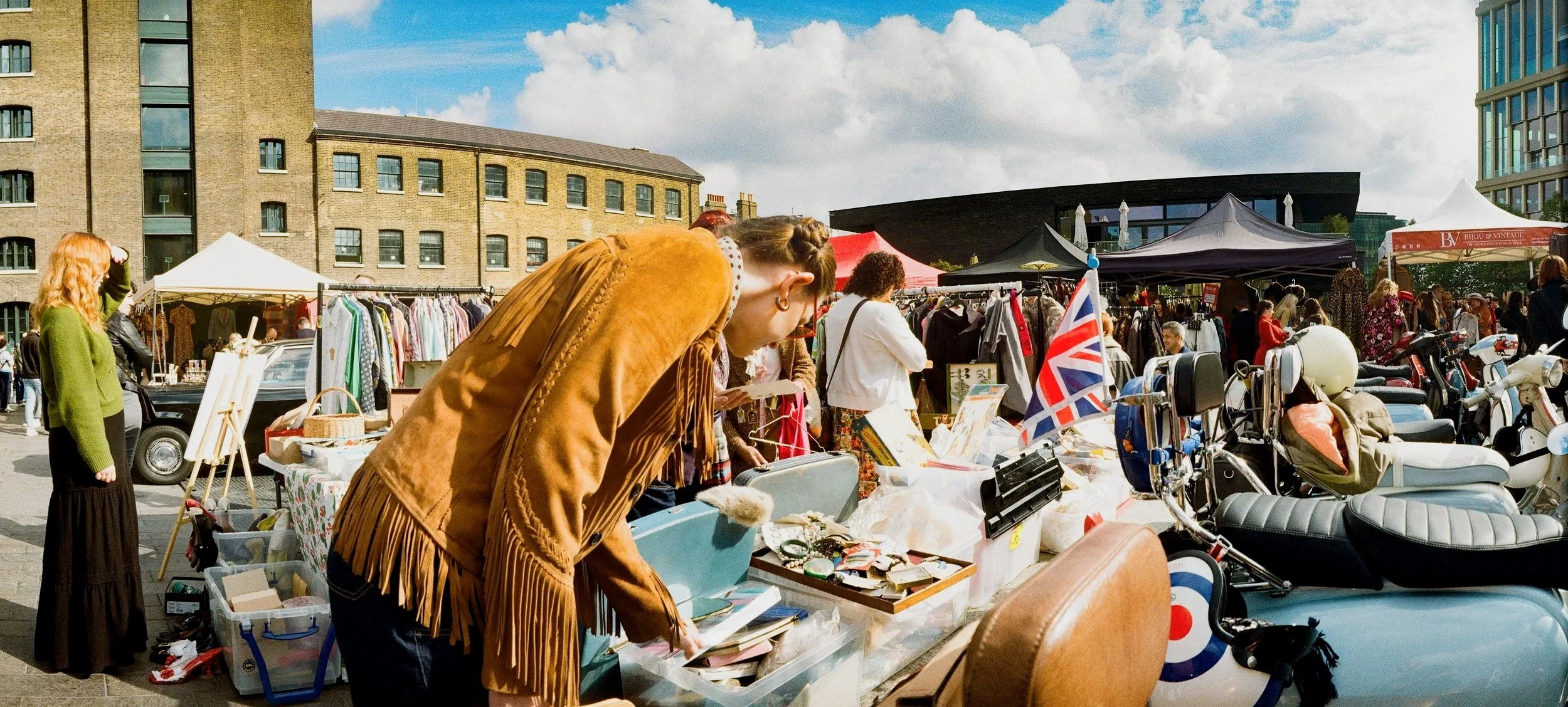 People browsing outdoor market stalls with clothing, accessories, and vintage items, motorcycles parked in front, and a UK flag, in an urban setting with modern and historic buildings under a partly cloudy sky.