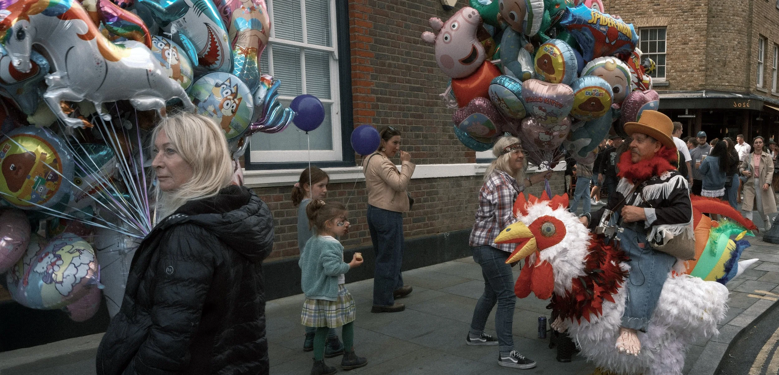 Street scene with people selling and holding colorful balloons, some with cartoon characters, outside a brick building. A woman is dressed in a chicken costume with a large white and red chicken head, interacting with another person.