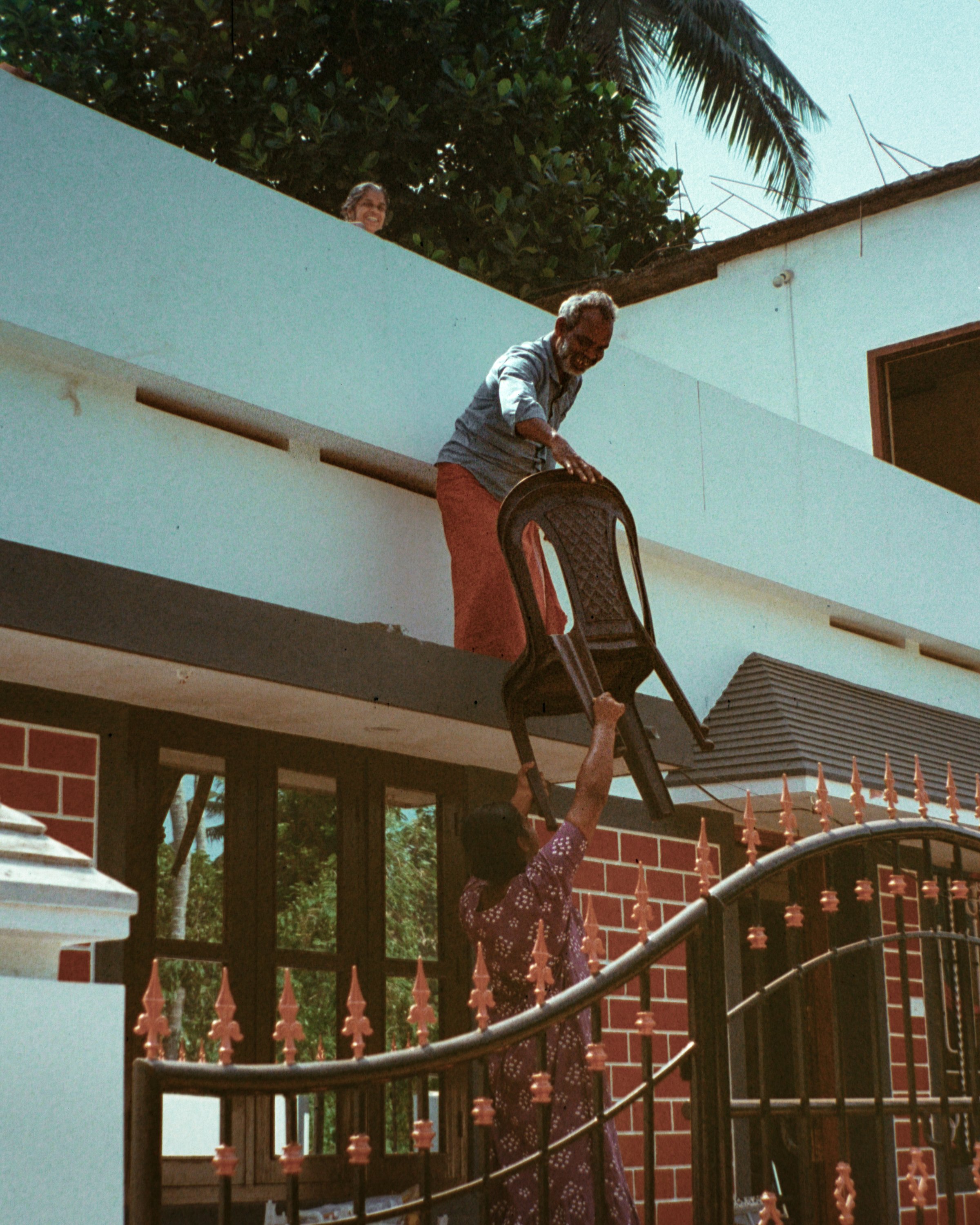 A person on the balcony giving a chair to another person below, with a woman on the balcony smiling.