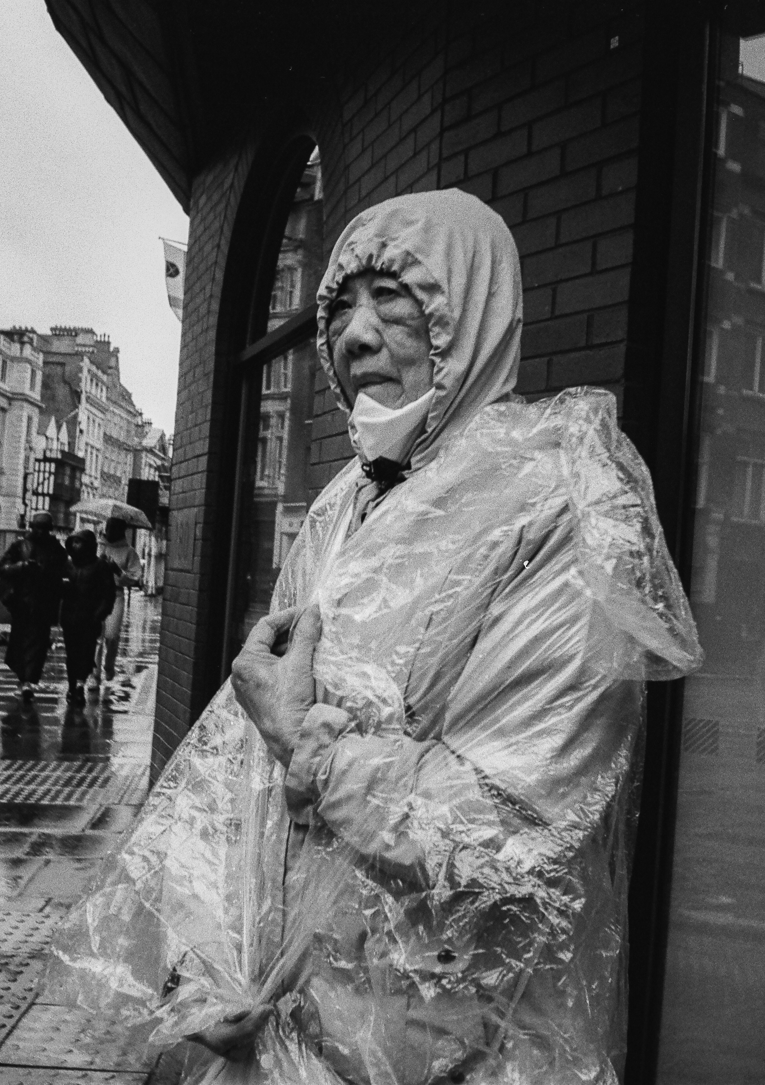 An elderly woman standing on a rain-soaked city sidewalk, wearing a hooded rain poncho and a face mask, looking pensively to the side in black and white.