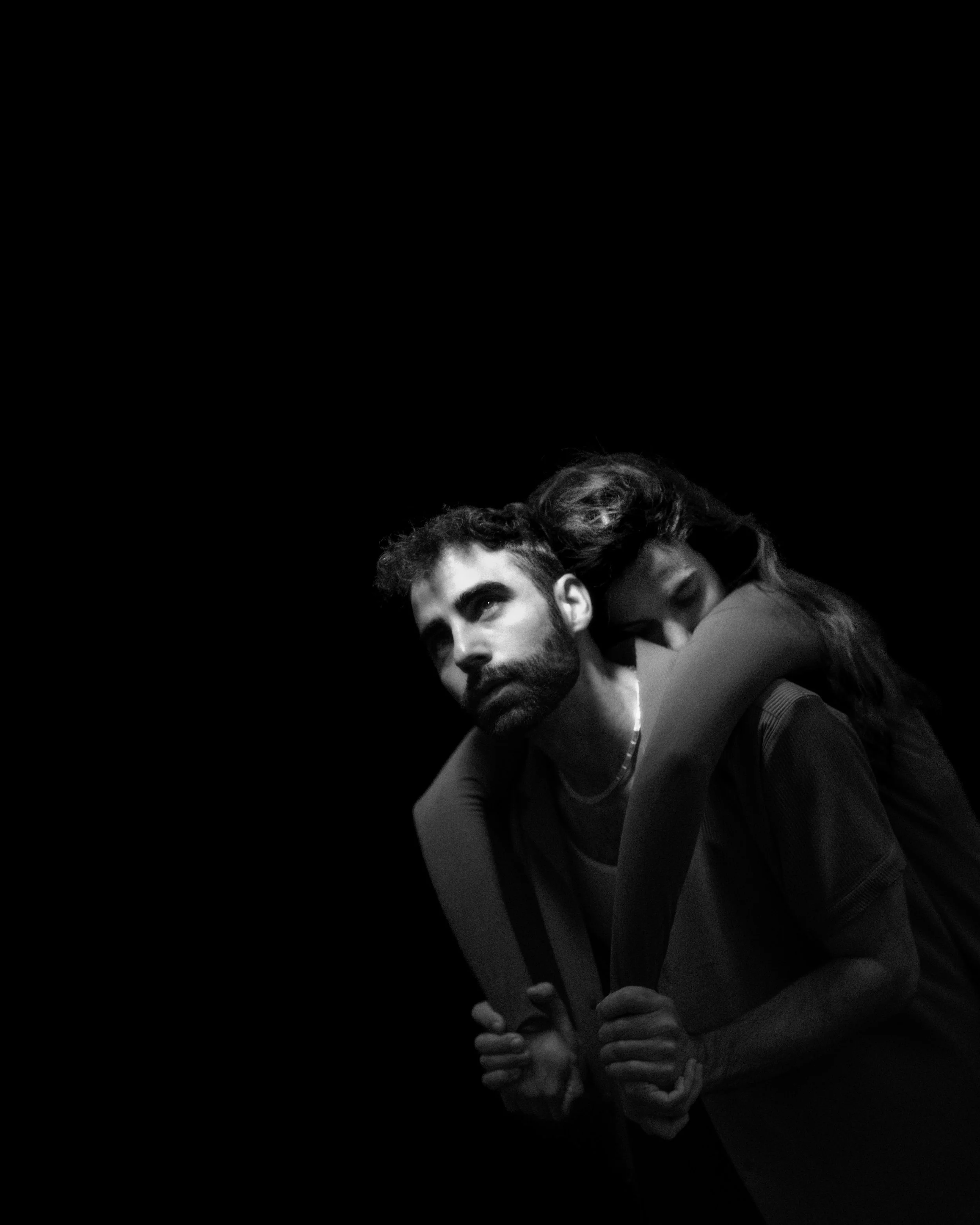 A black and white photograph of a young couple; the man has a beard and short hair, looking pensively, while the woman with long wavy hair embraces him from behind, resting her head on his shoulder.