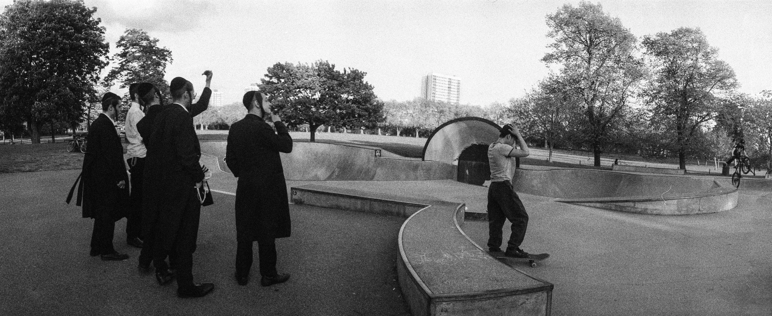 A black and white photo of a skatepark with five men and one boy, some in formal attire, pointing and watching a boy skateboarding near a concrete ramp, with trees and a building in the background.