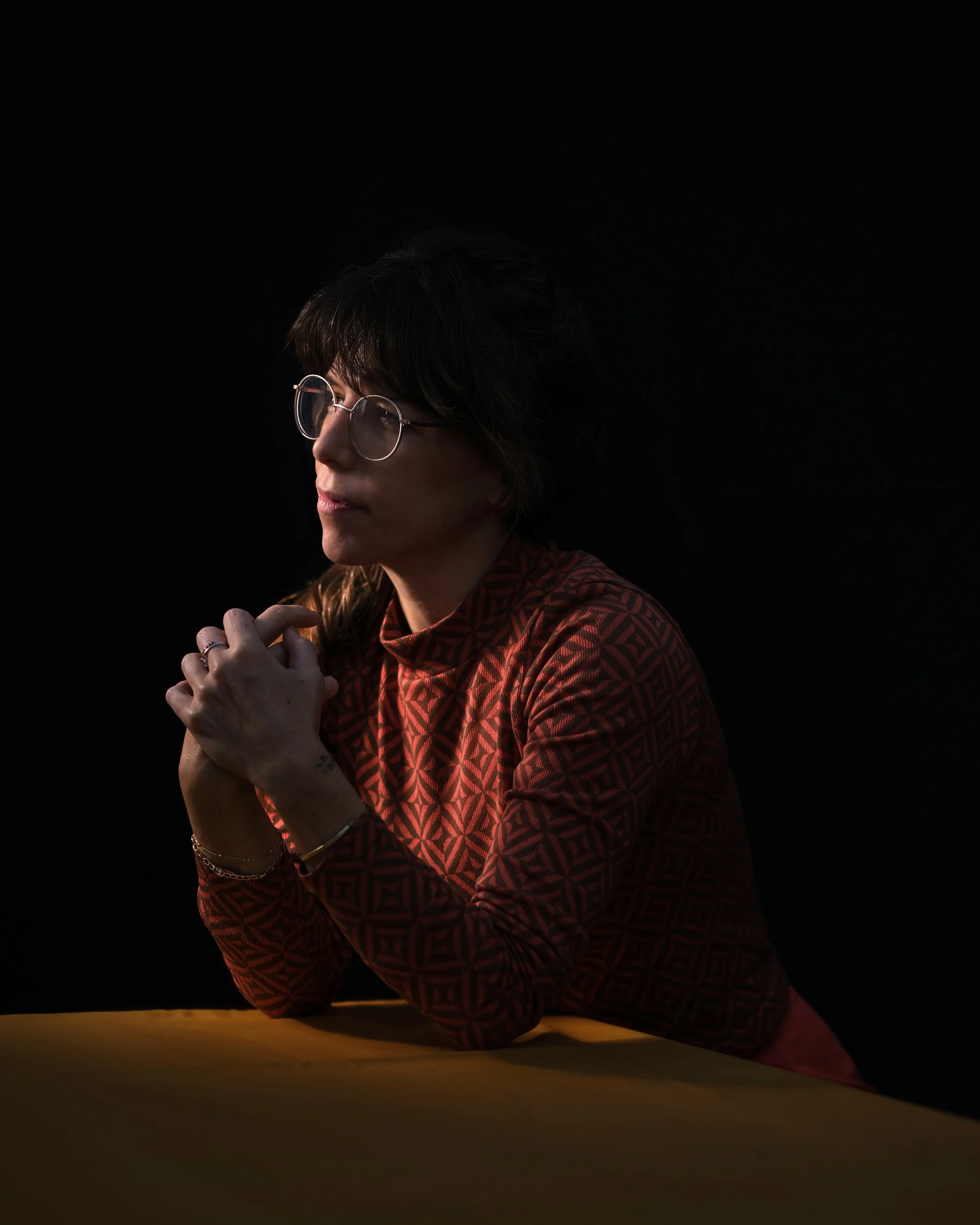 A woman with glasses in a red patterned top, sitting at a table with her hands clasped, against a dark background.