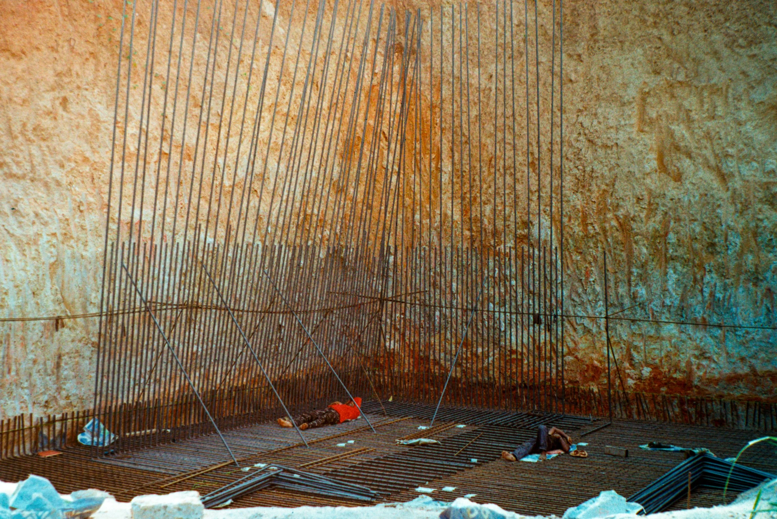Two people lying on the ground at a construction site with steel reinforcement bars and a rough textured wall in the background.